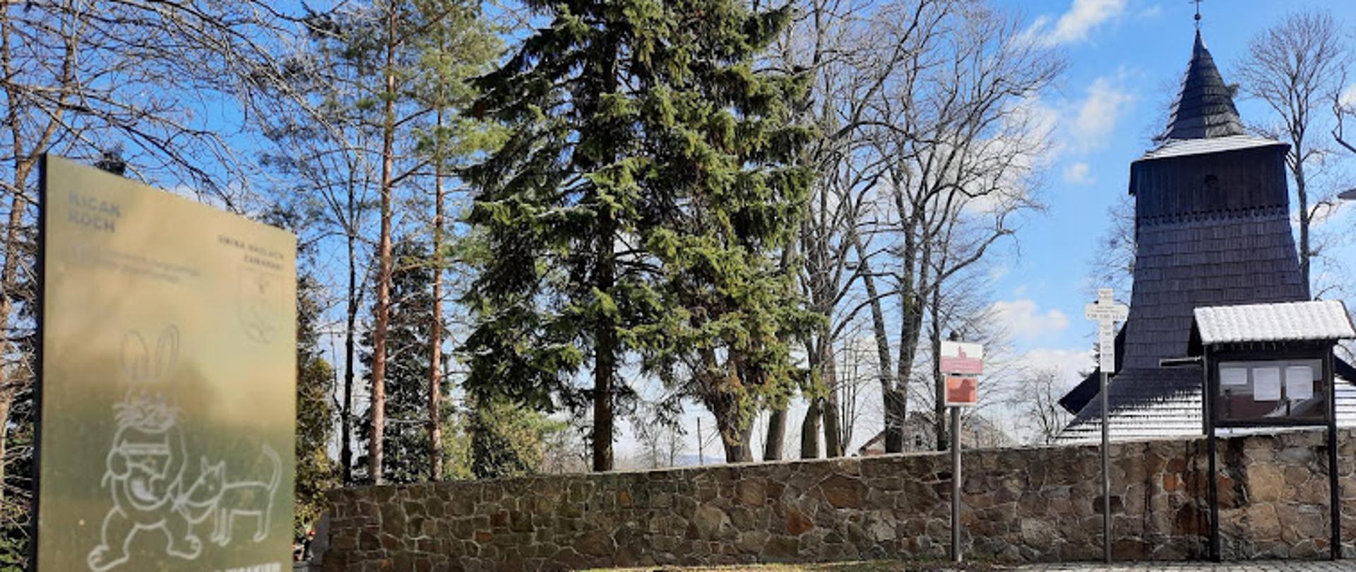 Na pierwszym planie słupek z tabliczką z Kicakiem. Na drugim planie zabytkowy kościół drewniany w Zamarskach