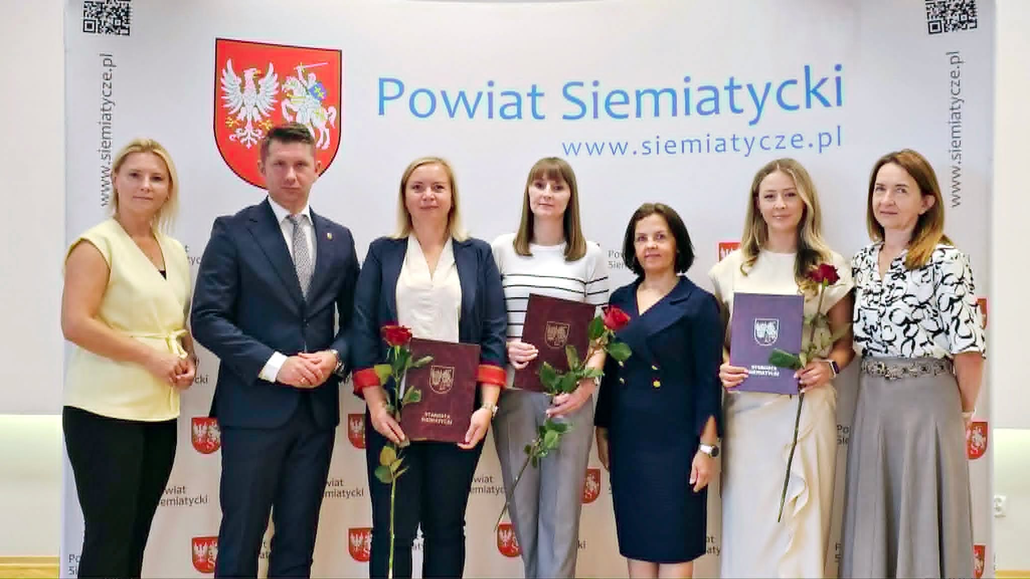 Grupa osób stojących razem przed banerem Powiatu Siemiatyckiego, trzymających certyfikaty i czerwone róże.