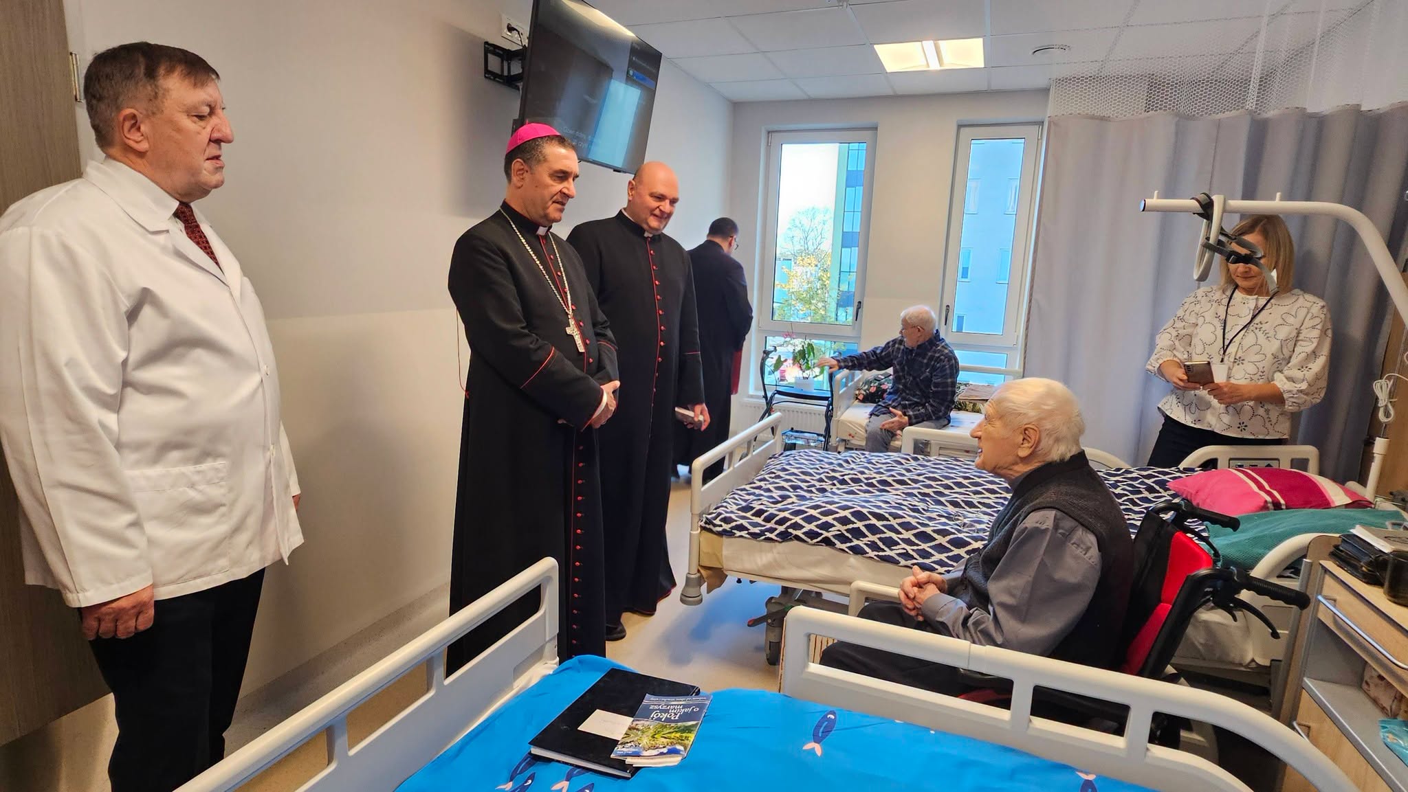 Grupa osób w sali szpitalnej, w tym dwóch duchownych i personel medyczny, rozmawiających z pacjentem leżącym w łóżku.