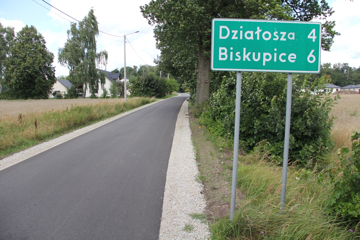 Nowy asfalt na drodze ciągnie się kilometrami. Z prawej znak drogowy, w tle pola i łąki