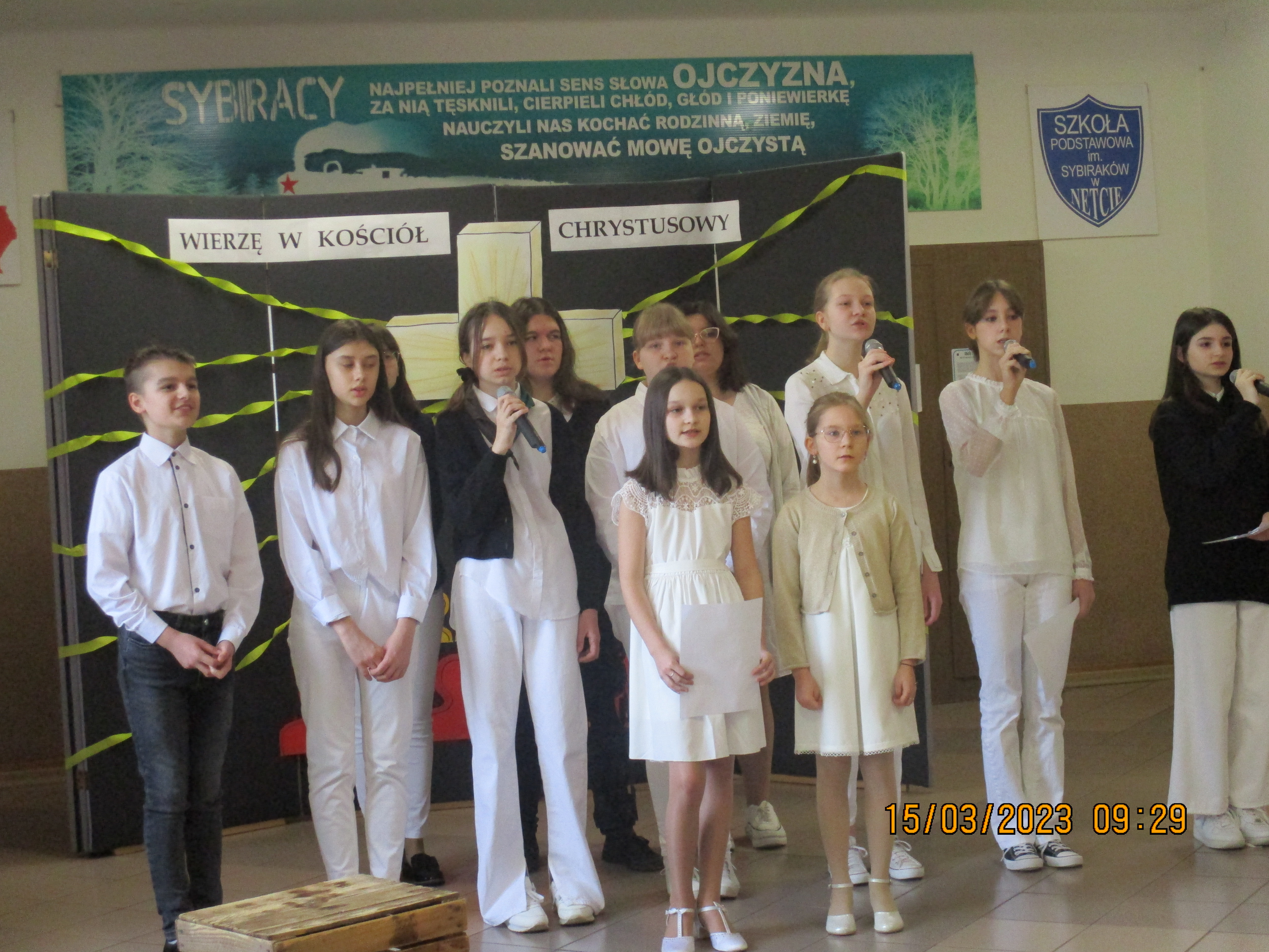 Grupa uczniów ubrana na biało śpiewa piosenkę. Za nimi, na dekoracji wisi napis: WIERZĘ W KOŚCIÓŁ CHRYSTUSOWY.