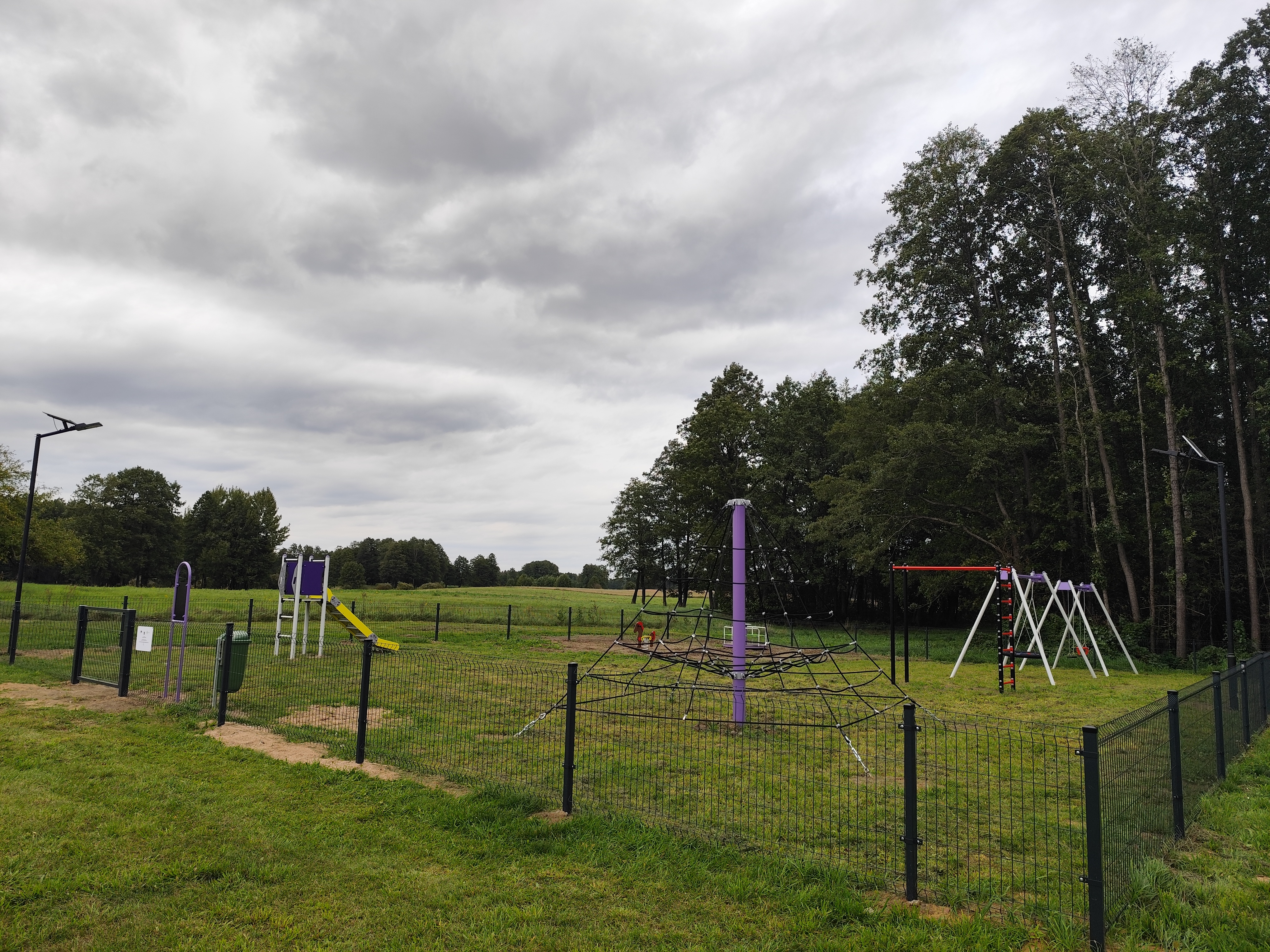 Zdjęcie przedstawia wiejski plac zabaw ogrodzony metalowym płotem. Widać na nim huśtawki, karuzelę, zjeżdżalnię oraz drabinki do wspinaczki.