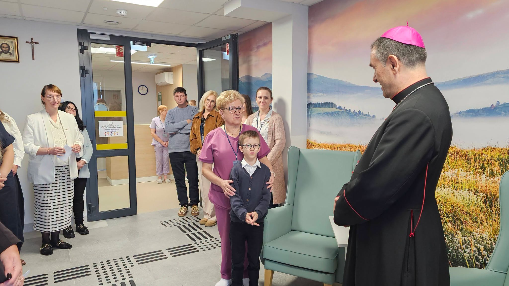 Grupa osób w pomieszczeniu z malowidłem ściennym przedstawiającym pejzaż; chłopiec stoi przed mężczyzną w stroju religijnym, podczas gdy inni obserwują tę scenę z daleka.