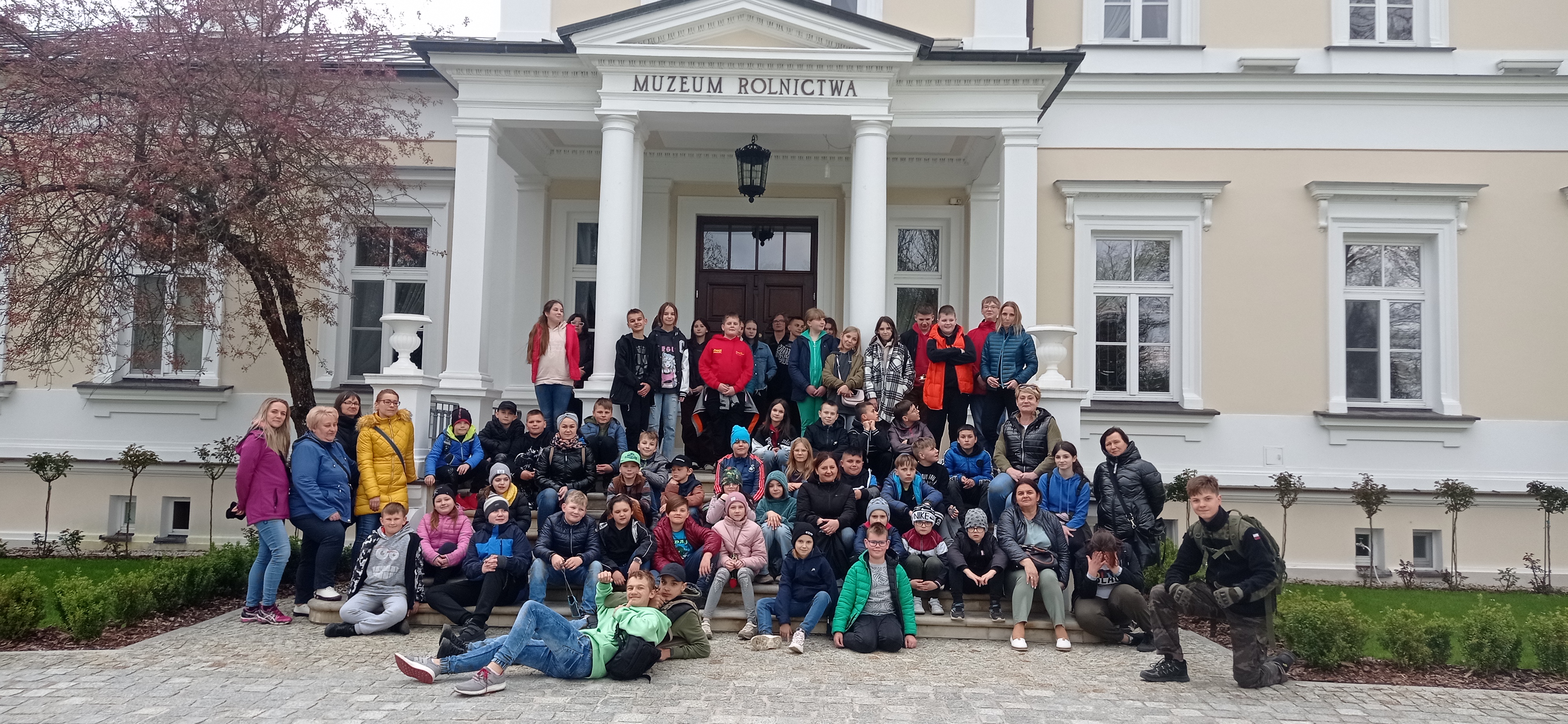 Grupowe zdjęcie wszystkich uczestników wycieczki pod dużym, białym budynkiem Muzeum Rolnictwa.