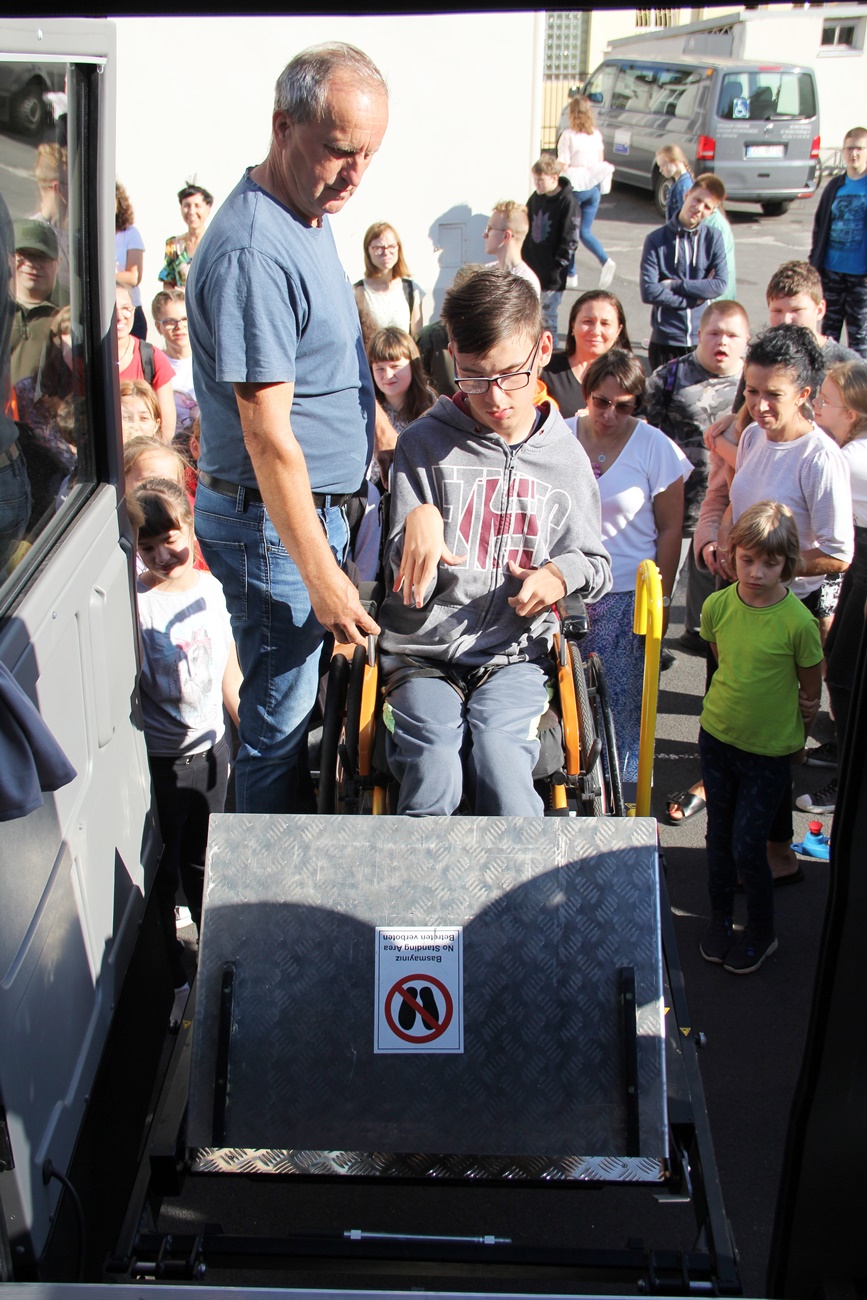Uczeń na wózku inwalidzkim wjeżdża do autobusu na windzie dla niepełnosprawnych. Winda umiejscowiona jest przy tylnym wejściu. Obok stoją uczniowie, którzy przyglądają się działaniu windy
