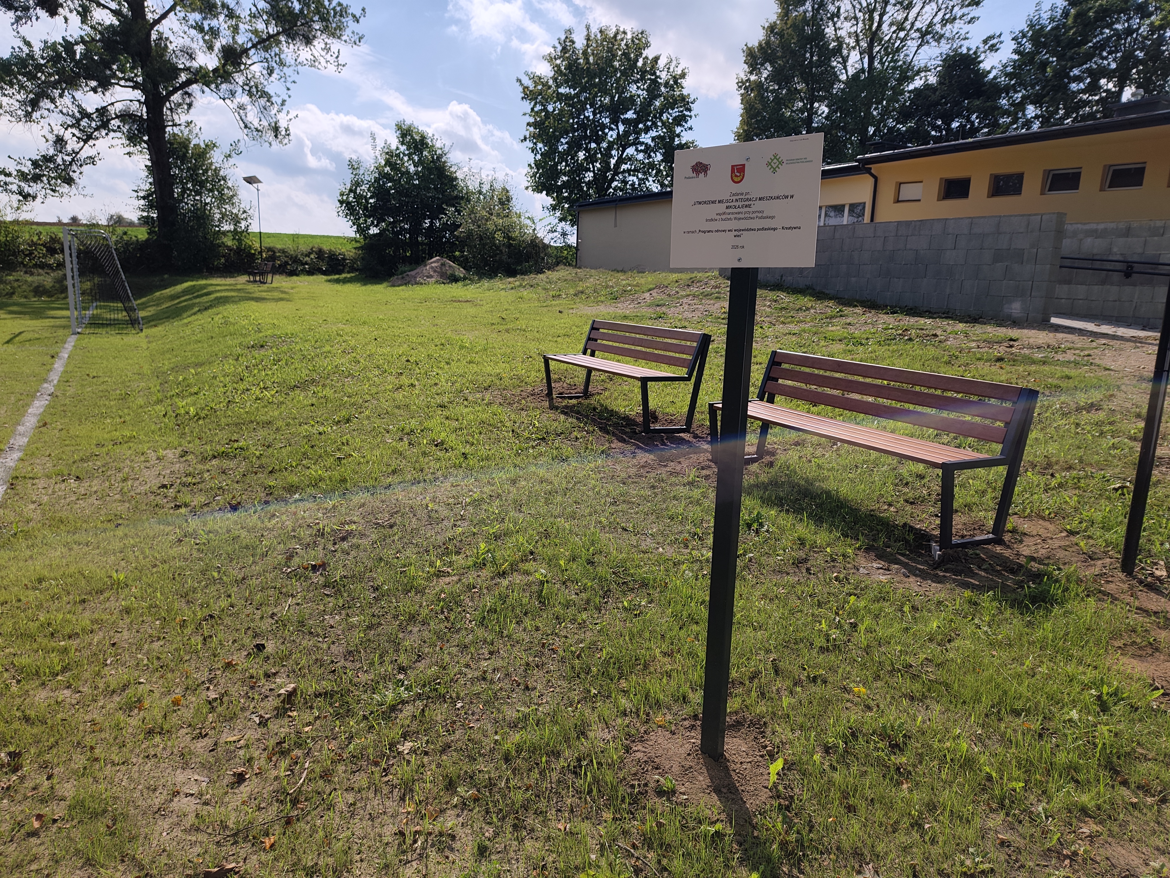 Trawiaste boisko piłkarskie z bramką i białymi liniami, obok którego stoją dwie ławki oraz tablica informacyjna. W tle znajduje się budynek, drzewa, lampa solarna i otwarte pola.