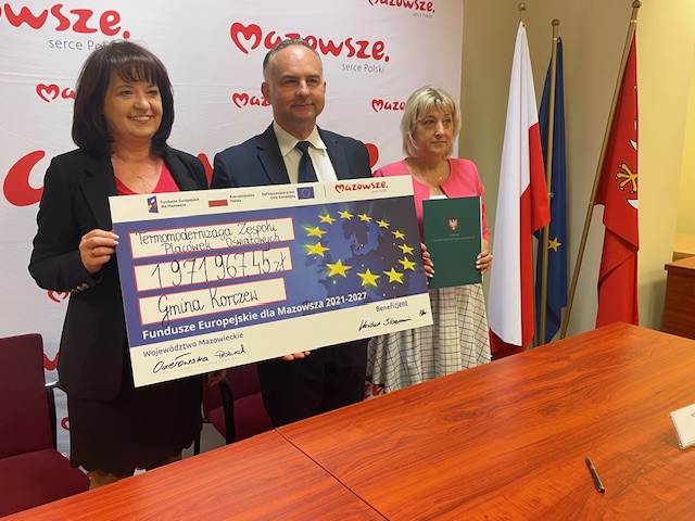 trzy osoby- dwie kobiety i mężczyzna stojące na tle białej tablicy z czerwonym napisem "Mazowsze. Dwie osoby trzymają czek z napisem "Termomodernizacja Zespołu Placówek Oświatowych 1 971 967,45 zł. Gmina Korczew. po prawek w kacie stojak z flagami: Polski, Województwa Mazowieckiego, Unii Europejskiej 