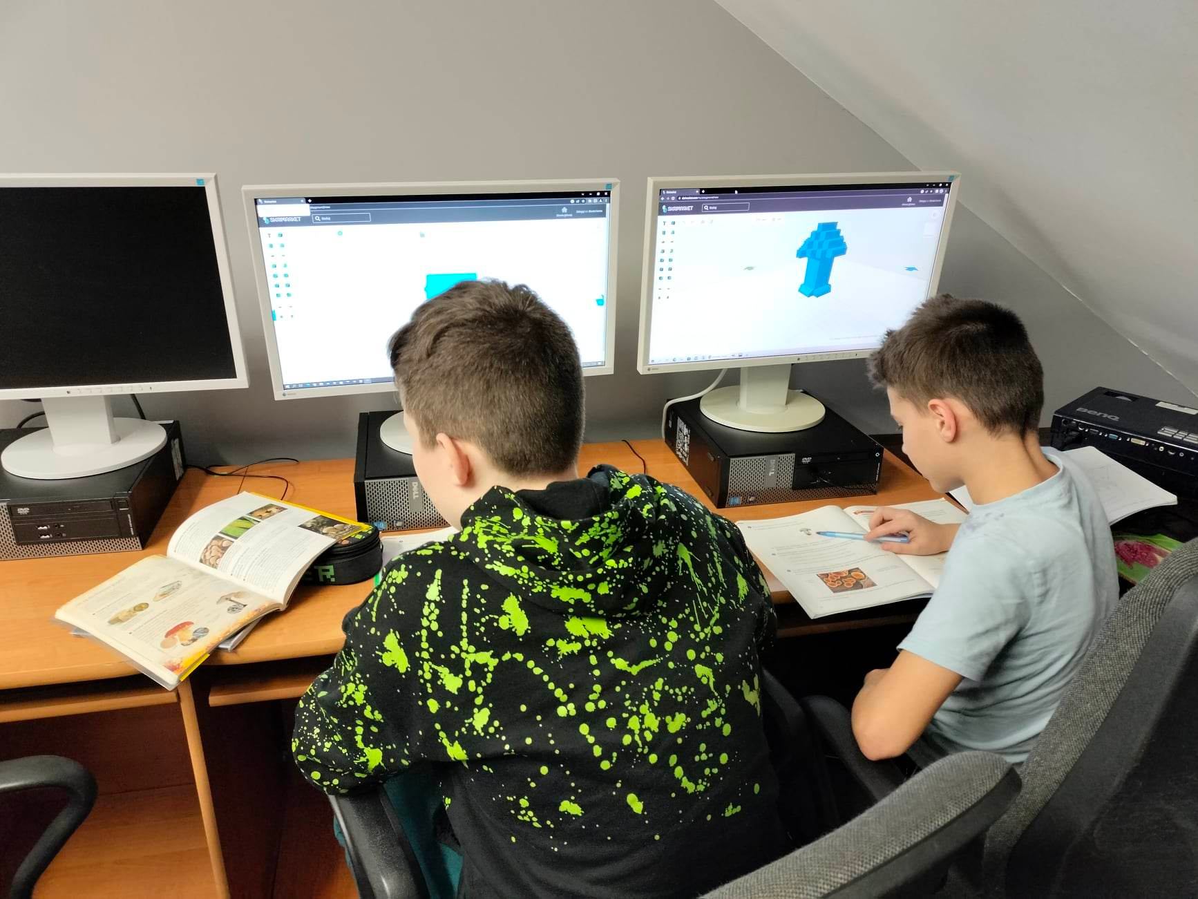 Dwóch uczniów przygląda się obrazkom umieszczonym w książkach. Obok stoją monitory z zaczętymi projektami modeli 3D.
