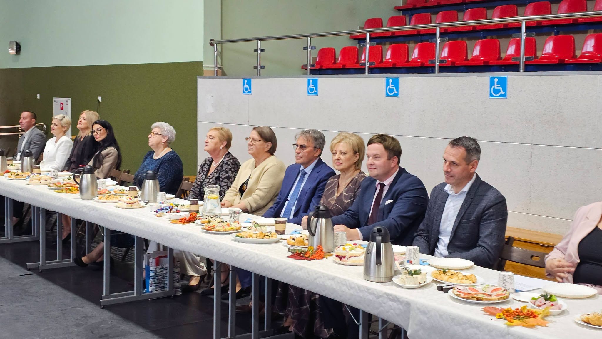 Długi stół nakryty jedzeniem i napojami w sali, przy którym po jednej stronie siedzi rząd osób.
