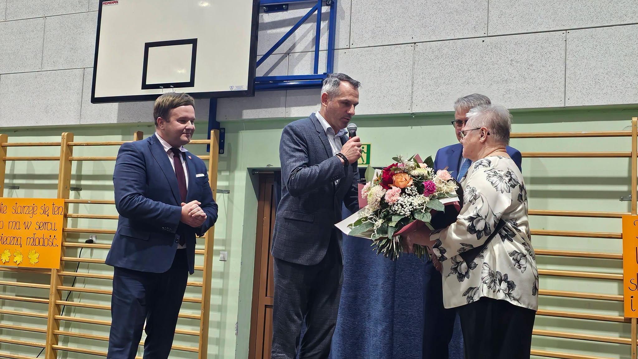 Grupa osób stojąca na scenie; jedna z nich wręcza bukiet kwiatów innej osobie, a dwie pozostałe przyglądają się tej scenie.