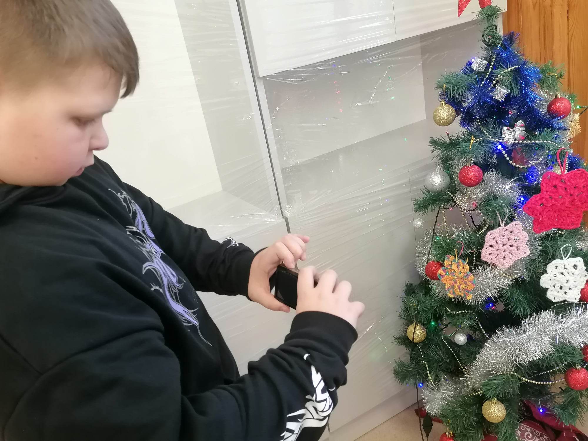 Chłopiec trzyma w dłoniach aparat i wykonuje zdjęcie choince przyozdobionej w kolorowe bombki zrobione z włóczki.