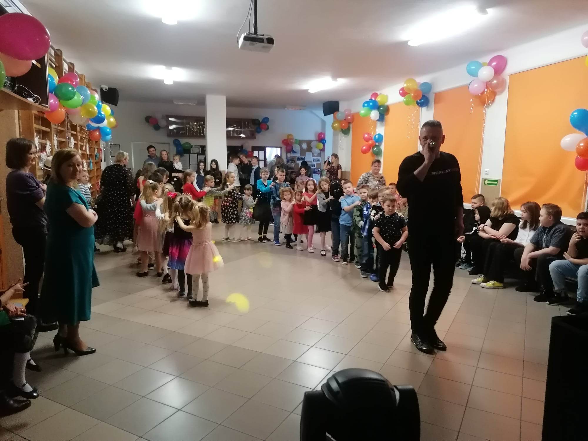 Mężczyzna ubrany w czarną bluzę trzyma w ręku mikrofon. Za nim ustawiony jest rząd dzieci w wieku przedszkolnym. Wokół nich znajdują się starsi uczniowie, rodzice i nauczyciele. Na ścianach wiszą kolorowe balony.