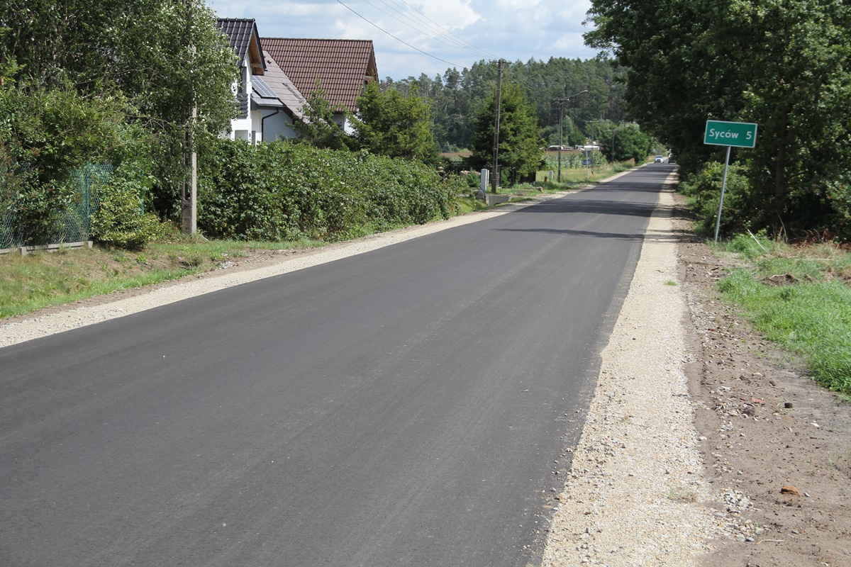 Nowy asfalt na drodze ciągnie się kilometrami. Po obu stronach białe pobocza, po prawej tablica informacyjna, w tle drzewa, łąki, lasy i domy