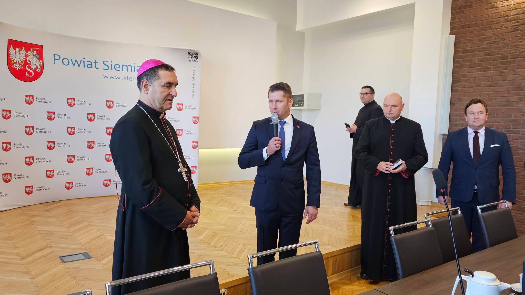 Grupa mężczyzn, w tym przywódca kościelny w czarnej szacie z różowym kołnierzem, stoi w sali konferencyjnej, a w tle widać ścianę prezentacyjną. Jeden z mężczyzn przemawia do mikrofonu, podczas gdy pozostali słuchają.