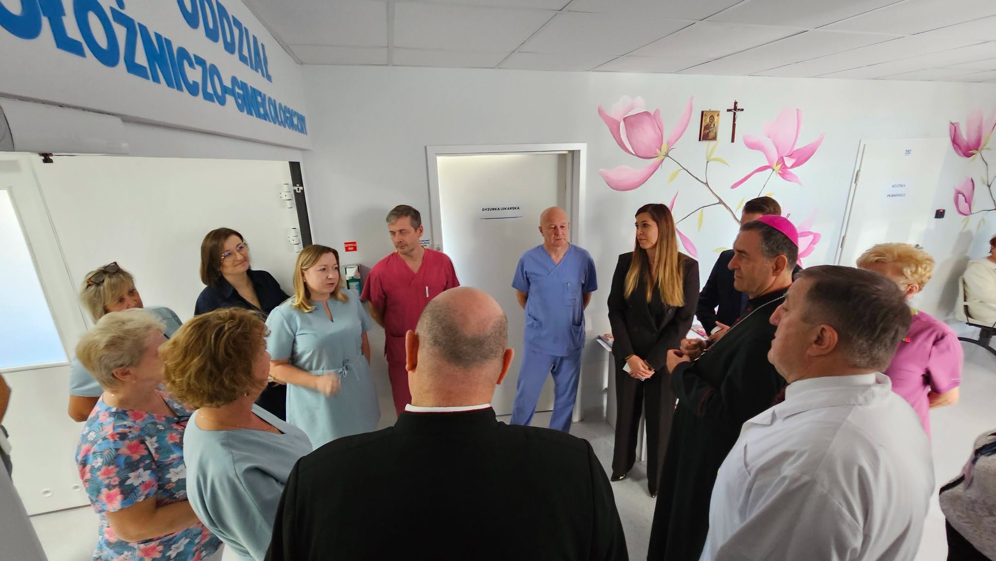 Grupa osób, w tym personel medyczny i duchowni, zgromadzonych w placówce opieki zdrowotnej, dyskutujących lub słuchających mówcy, przy ścianach ozdobionych kompozycjami kwiatowymi.