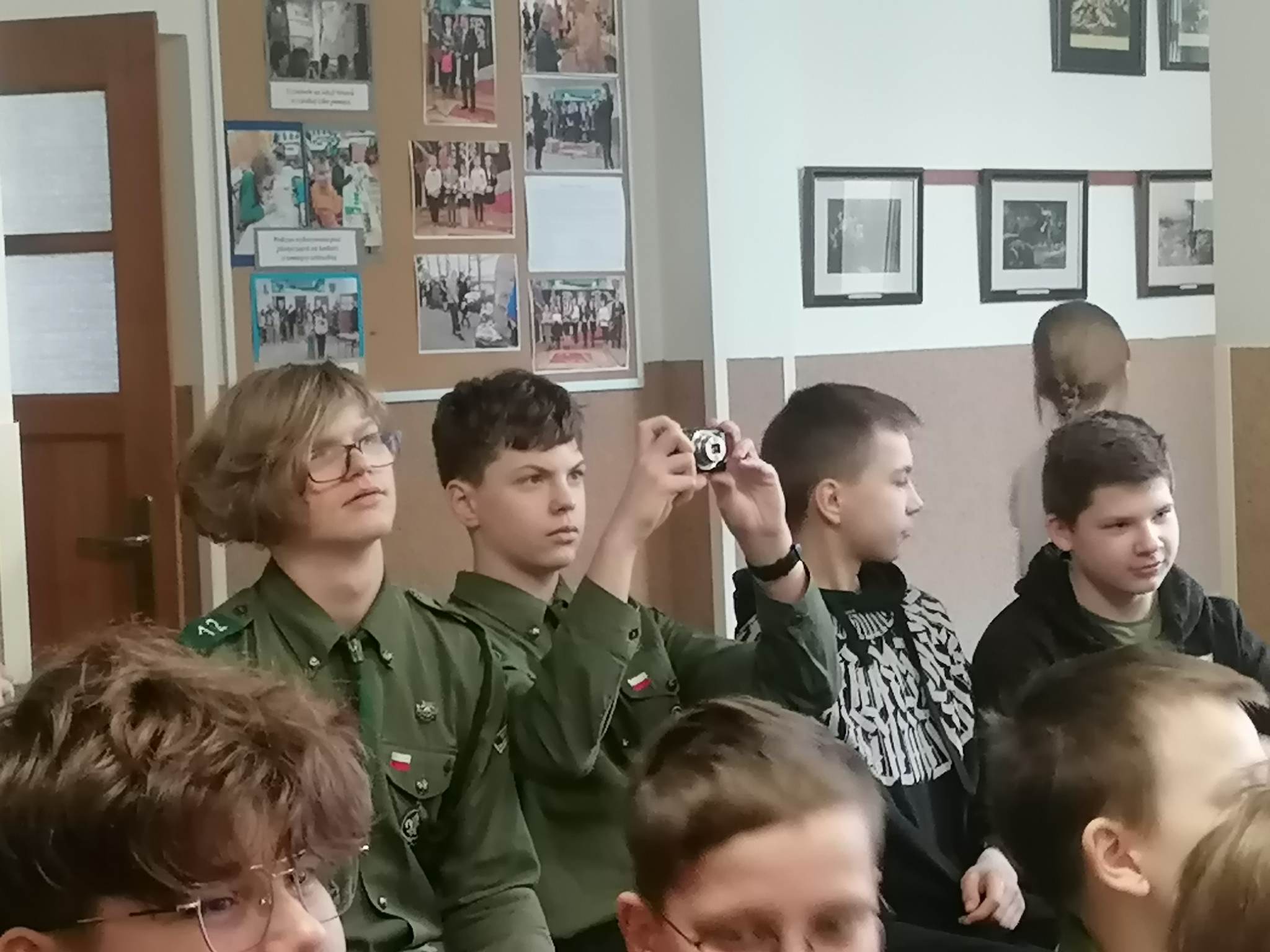 Czworo uczniów siedzi na krzesłach. Jeden z nich wykonuje zdjęcie aparatem cyfrowym.