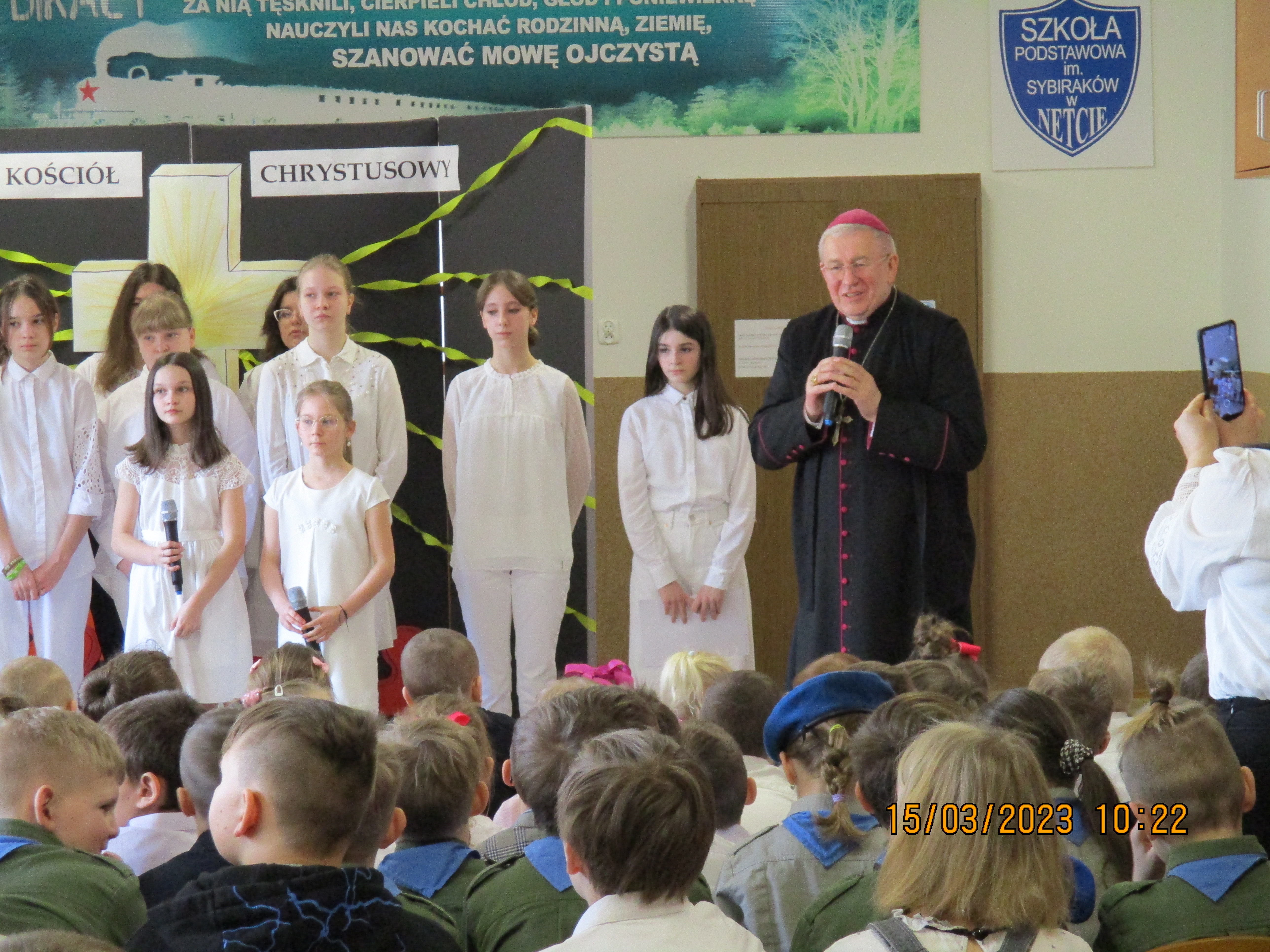 Na pierwszym planie stoi Biskup Ełcki - Jerzy Mazur. Duchowny trzyma w dłoniach mikrofon przez który przemawia do zgromadzonych uczniów. Za biskupem stoją dzieci ubrane na biało. Przed duchownym siedzą w ławkach uczniowie ubrani w harcerskie mundury.