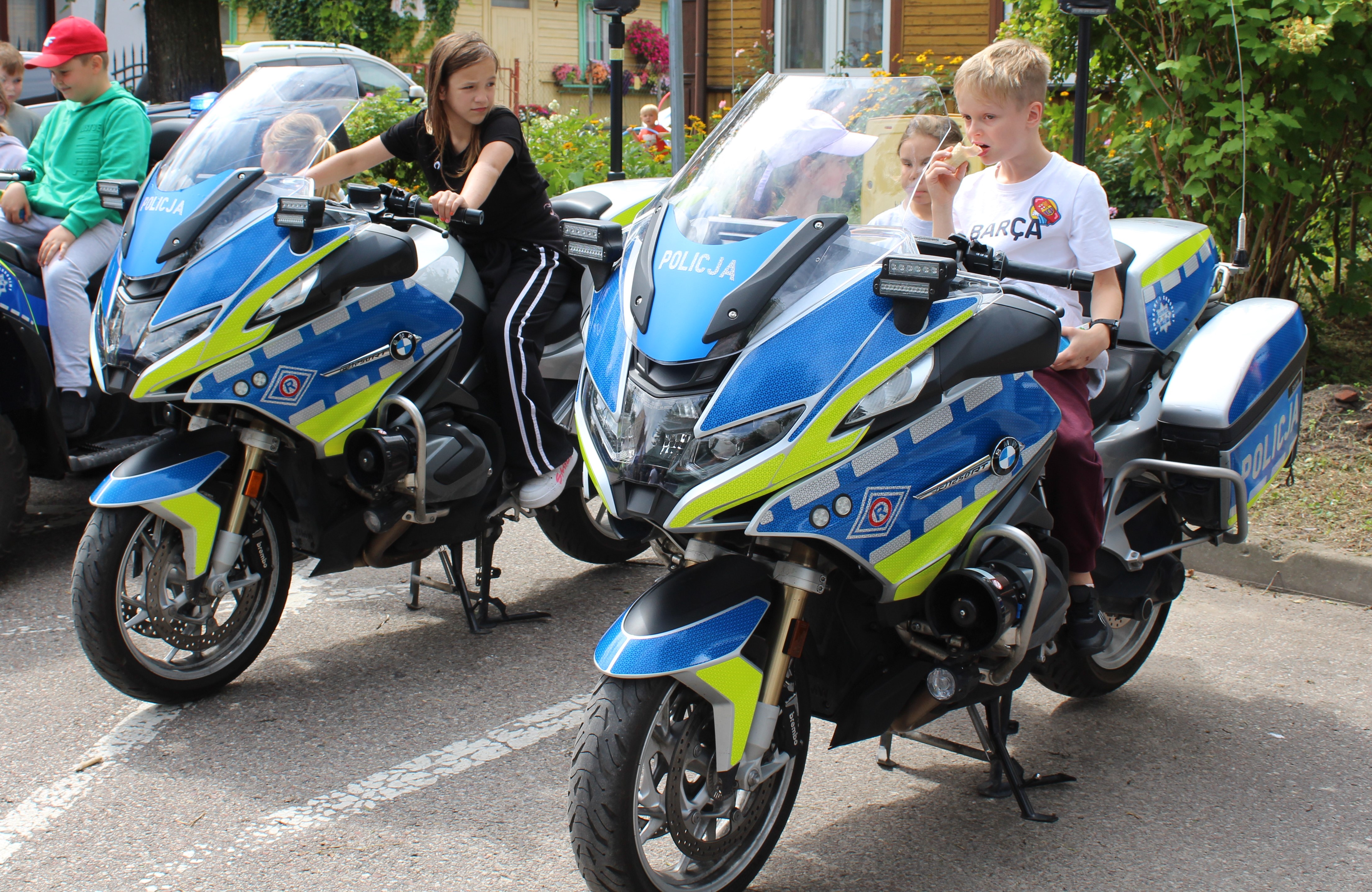 Dwoje dzieci siedzących na policyjnych motocyklach; jedno z nich przybiera poważną pozę, podczas gdy drugie wydaje się być pogrążone w głębokiej zadumie. Motocykle są pomalowane na jaskrawo niebieski i zielony kolor, z napisem „POLICJA”.