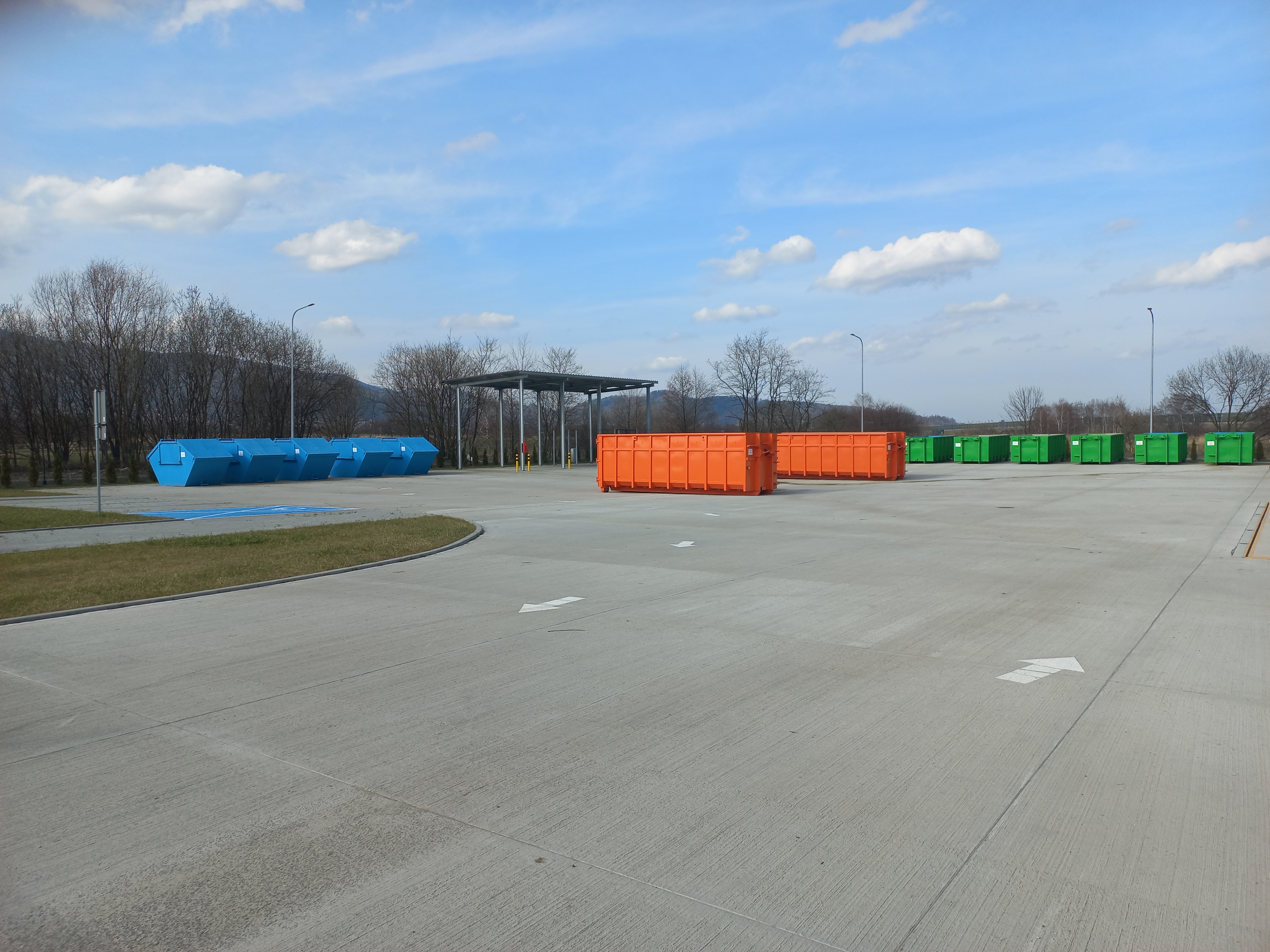 Grafika przedstawia betonowy plac z elementami zieleni, na którym ustawione są kontenery na odpady w różnych kolorach oraz, w oddali, wiata. W tle widoczne drzewa i góry.