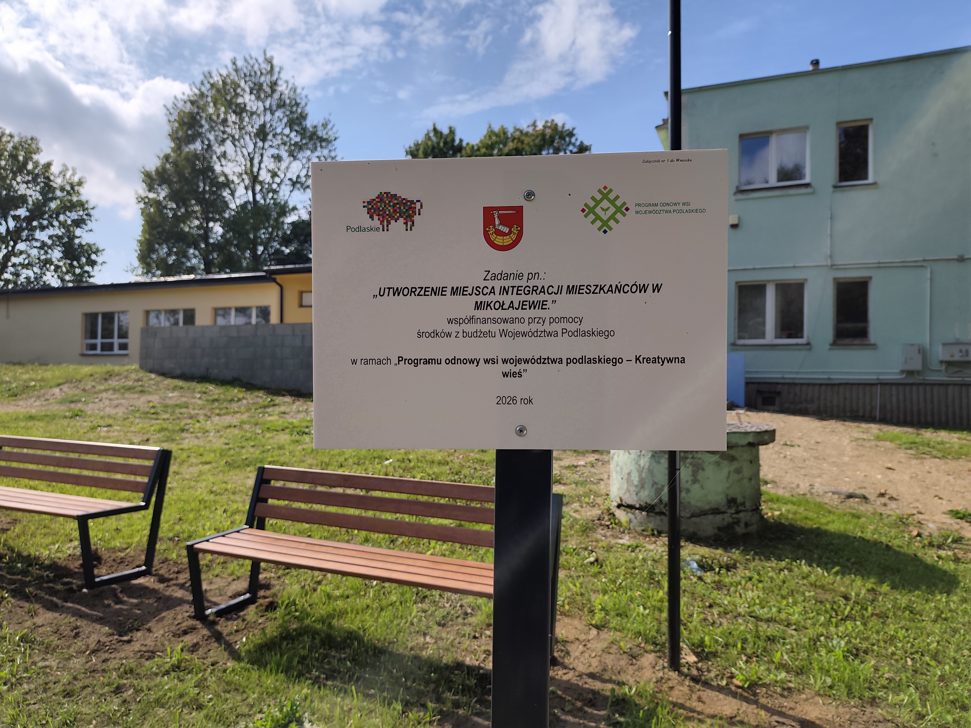 Tablica informacyjna przy dwóch ławkach na terenie rekreacyjnym. Na tablicy widnieje informacja o zadaniu „Utworzenie miejsca integracji mieszkańców w Mikołajewie”, współfinansowanym ze środków budżetu Województwa Podlaskiego w ramach „Programu odnowy wsi województwa podlaskiego – Kreatywna wieś” w 2026 roku. W tle znajduje się budynek oraz trawnik