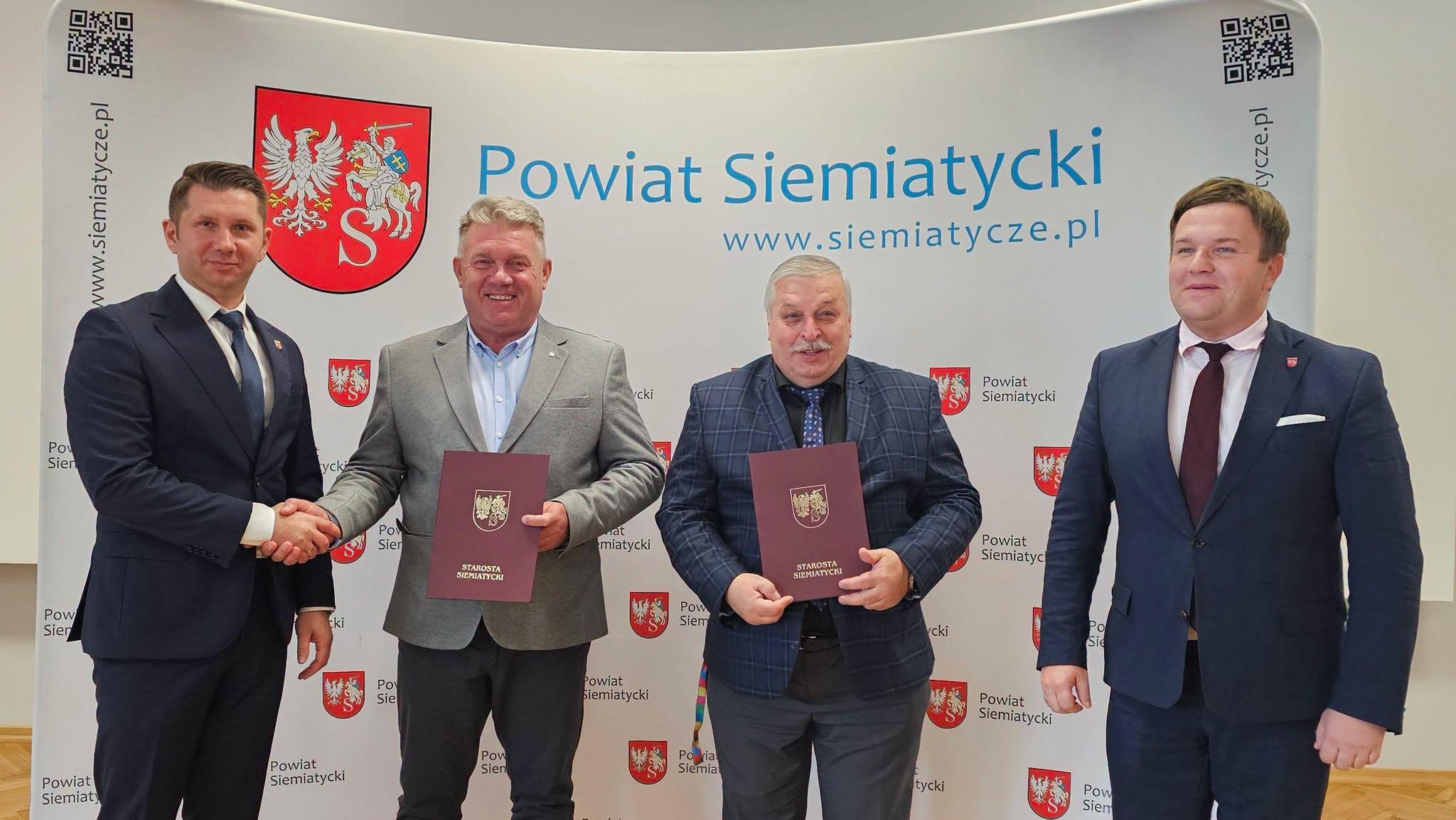 Czterech mężczyzn stoi przed transparentem z napisem „Powiat Siemiatycki”. Trzymają w rękach teczki i wygląda na to, że uczestniczą w uroczystym podpisaniu umowy lub ceremonii podpisania dokumentu.