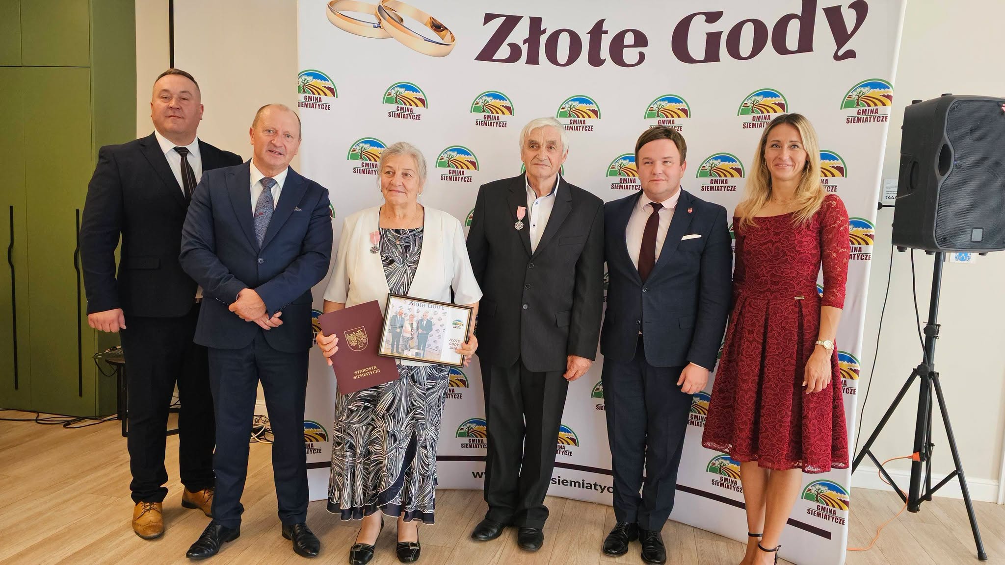 Grupa sześciu osób stoi przed transparentem z napisem „Złote Gody”. Dwie osoby trzymają dyplom. Są ubrane w stroje galowe.