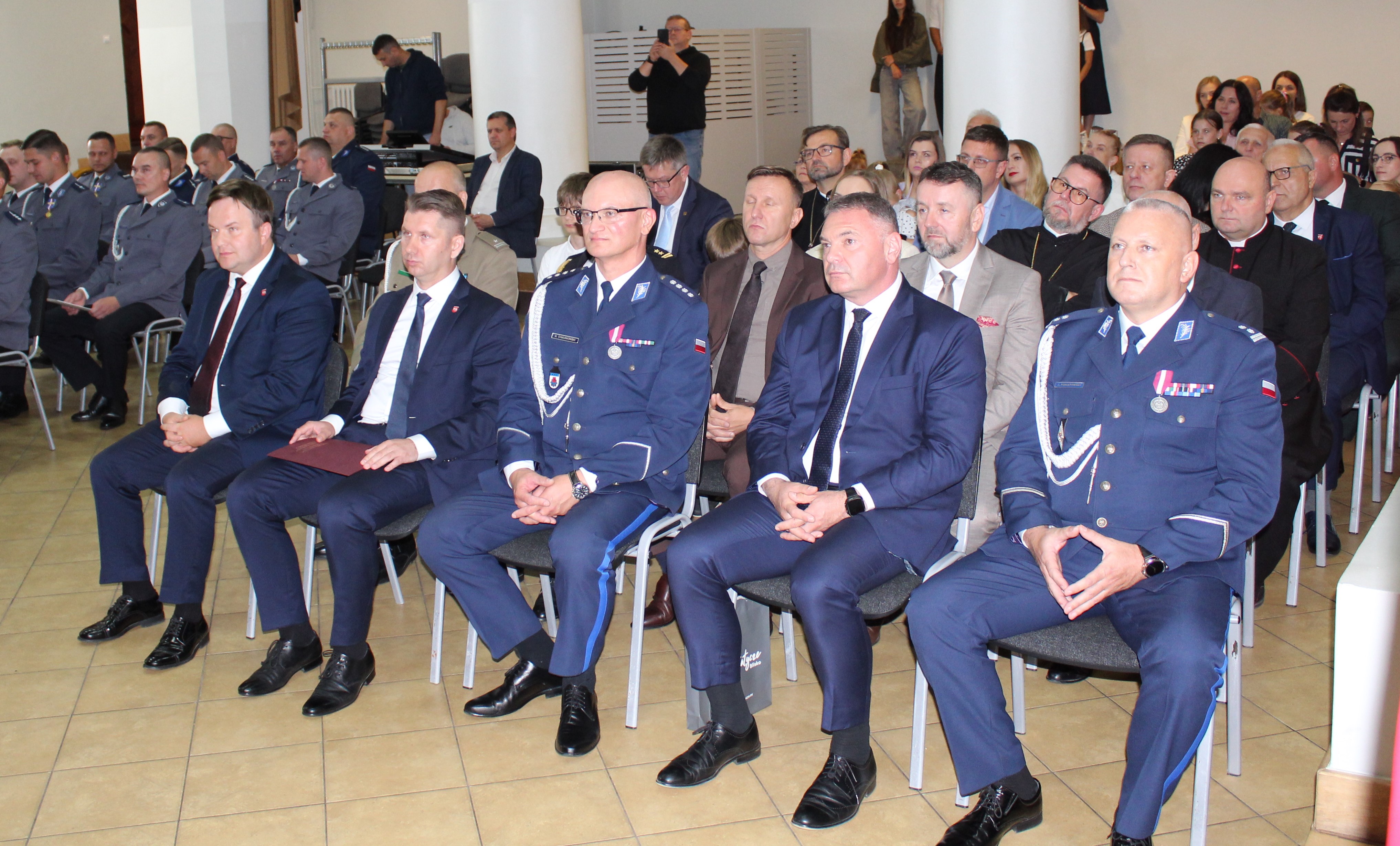Grupa mężczyzn w garniturach i mundurach siedzących w rzędach, uczestniczących w wydarzeniu.