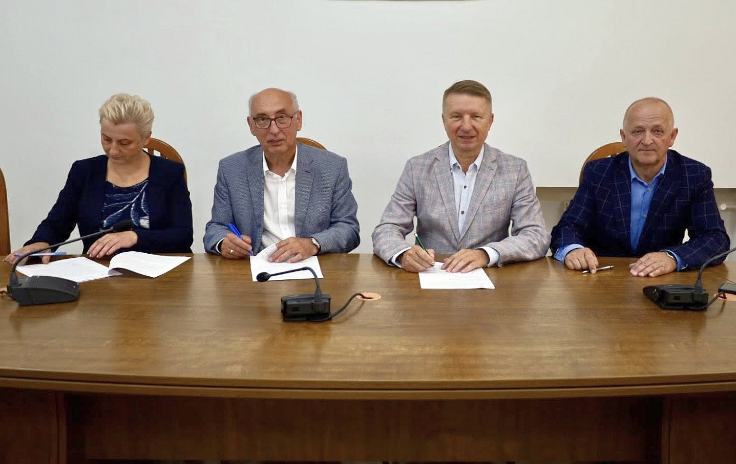 Na zdjęciu widzimy cztery osoby siedzące przy drewnianym stole konferencyjnym, podpisujące dokumenty. Wszyscy są elegancko ubrani, mężczyźni mają na sobie marynarki, a kobieta (pierwsza z lewej) nosi granatowy żakiet. Przed każdą z osób znajduje się mikrofon oraz dokumenty do podpisu. Tło jest neutralne, białe, co sugeruje, że zdjęcie wykonano w sali urzędowej lub podczas oficjalnego spotkania. Atmosfera jest formalna, a osoby na zdjęciu sprawiają wrażenie zaangażowanych w ważne ustalenia lub podpisanie umowy.
