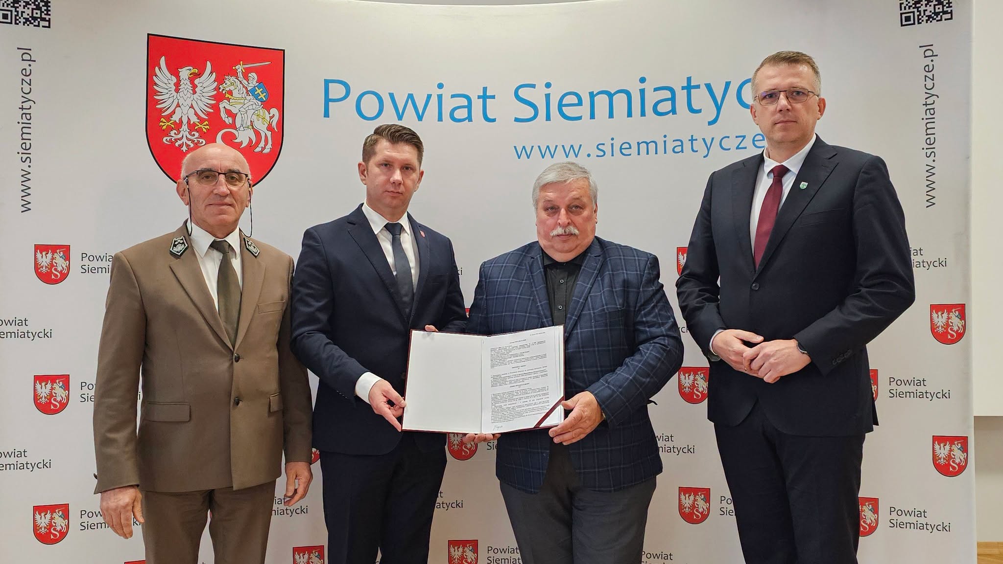 Grupa czterech mężczyzn stoi razem przed transparentem z napisem „Powiat Siemiatycki”. Jeden z mężczyzn trzyma w ręku dokument.