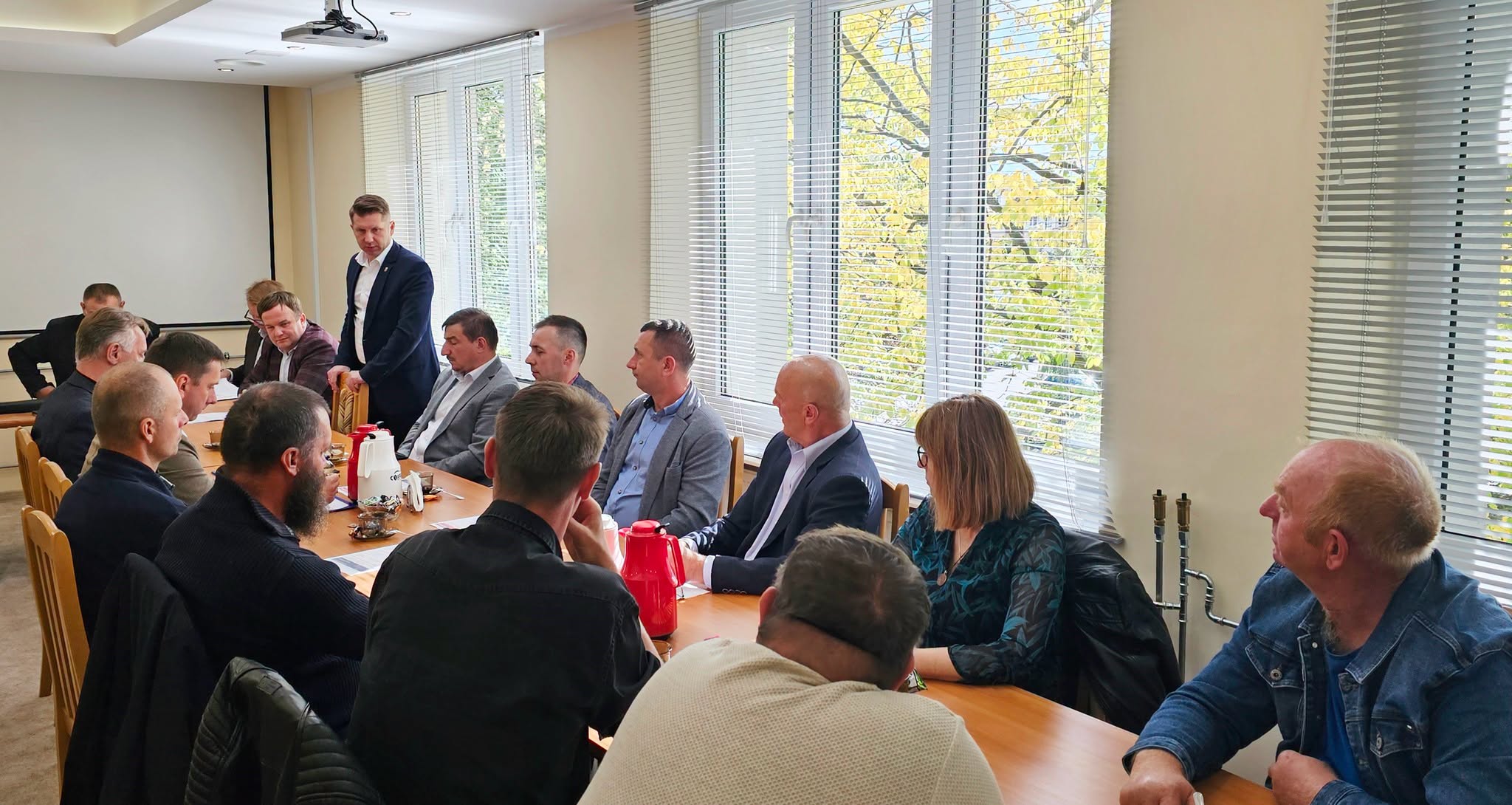 Grupa osób siedzących przy stole konferencyjnym, prowadząca dyskusję z kilkoma osobami stojącymi i przedstawiającymi prezentację. Duże okna w tle wpuszczają naturalne światło.
