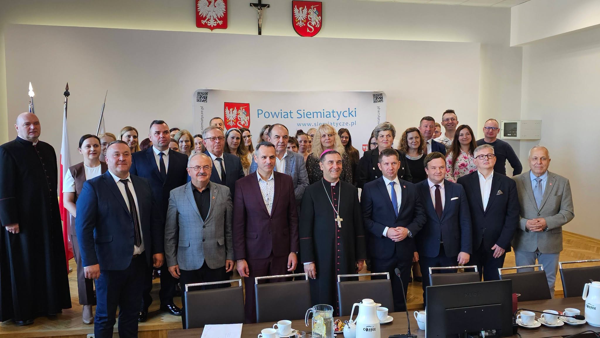 Duża grupa osób, w tym mężczyźni i kobiety w strojach galowych, stoi razem do zdjęcia w sali konferencyjnej, a w tle widać flagi i transparenty.