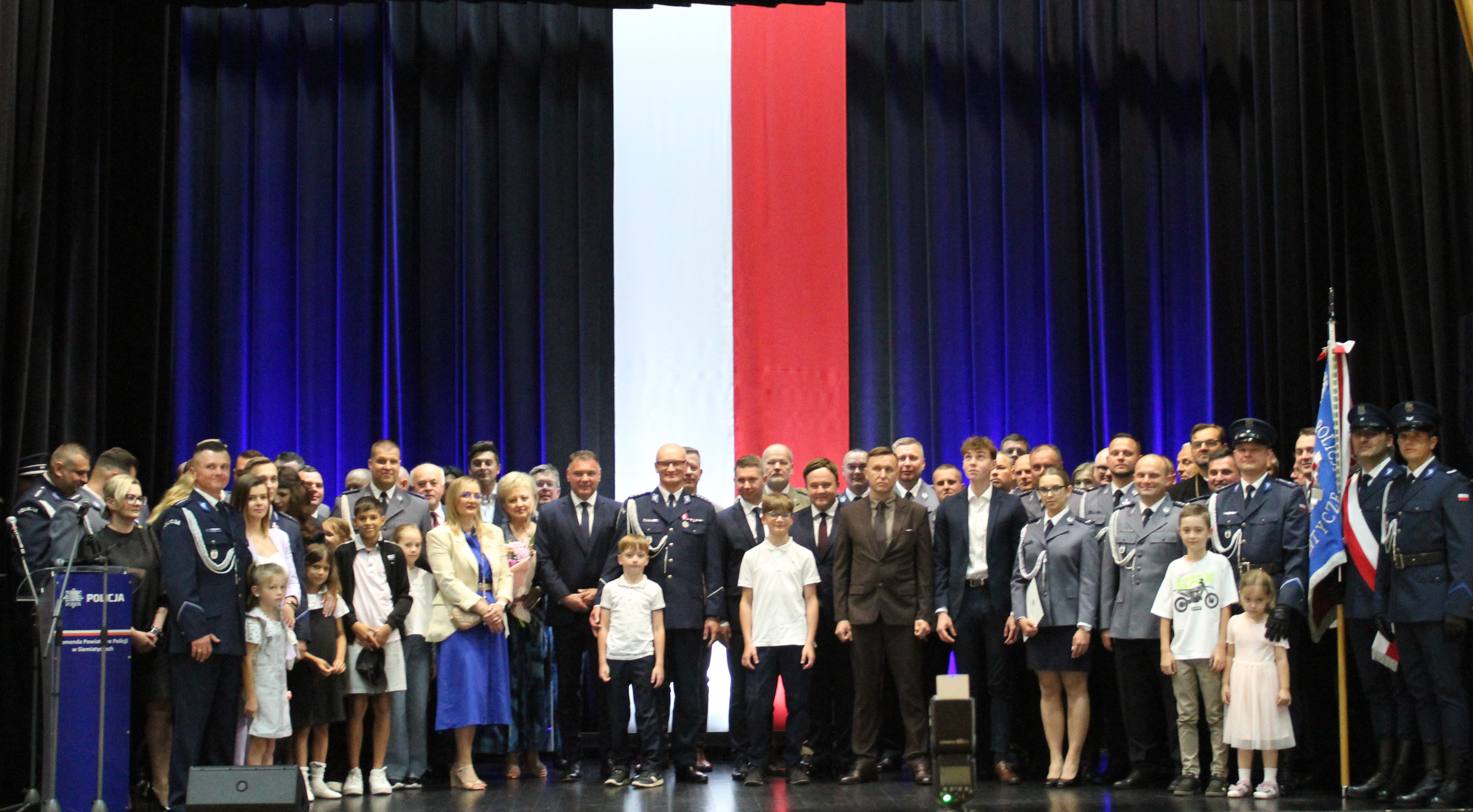 Duża grupa osób, w tym funkcjonariusze w mundurach i cywile, zgromadzonych na scenie przed dużym pionowym tłem w kolorach czerwonym i białym, z małą polską flagą po jednej stronie.
