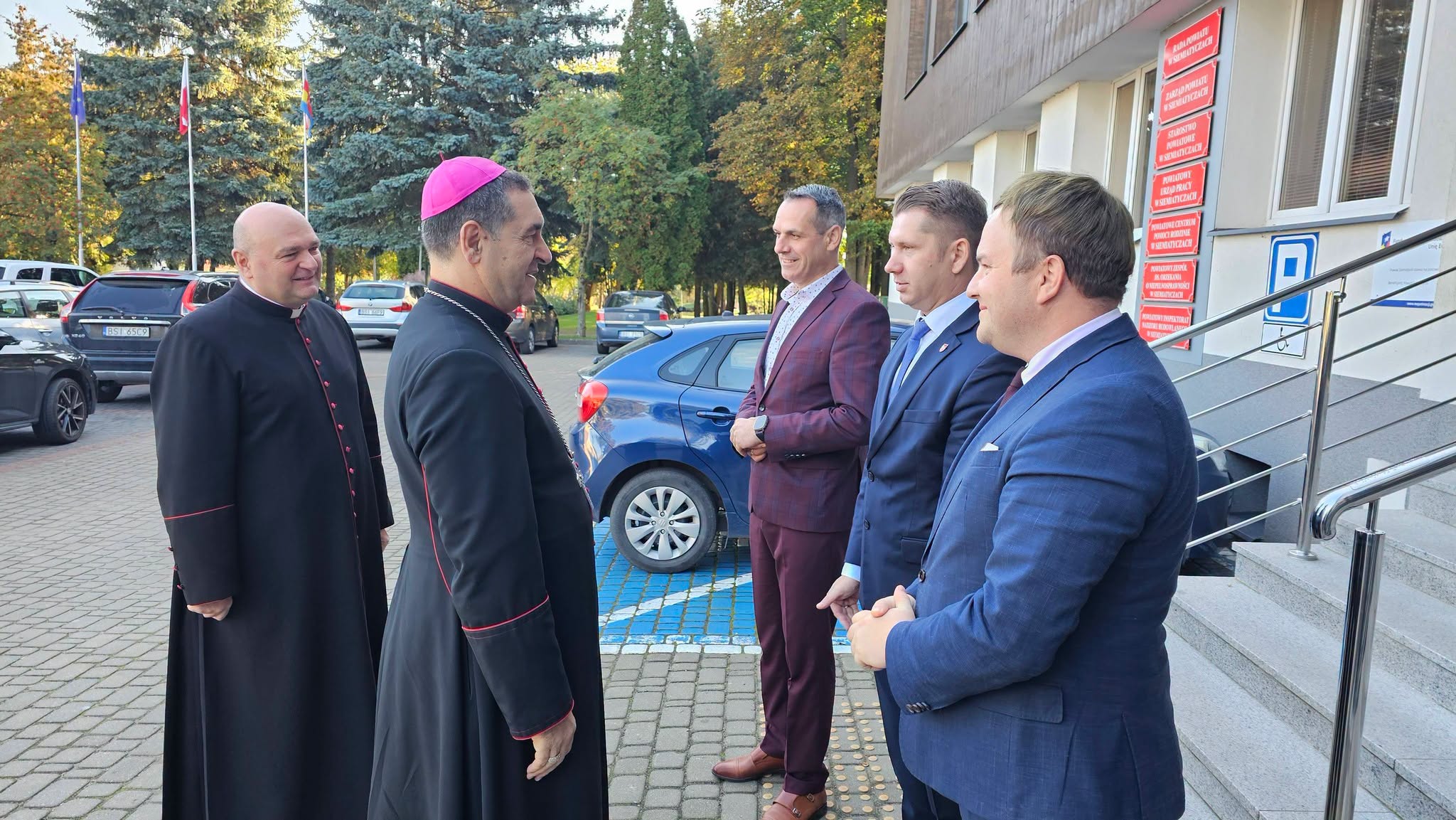 Grupa mężczyzn w strojach galowych rozmawia przed budynkiem, a w pobliżu stoi zaparkowany samochód.