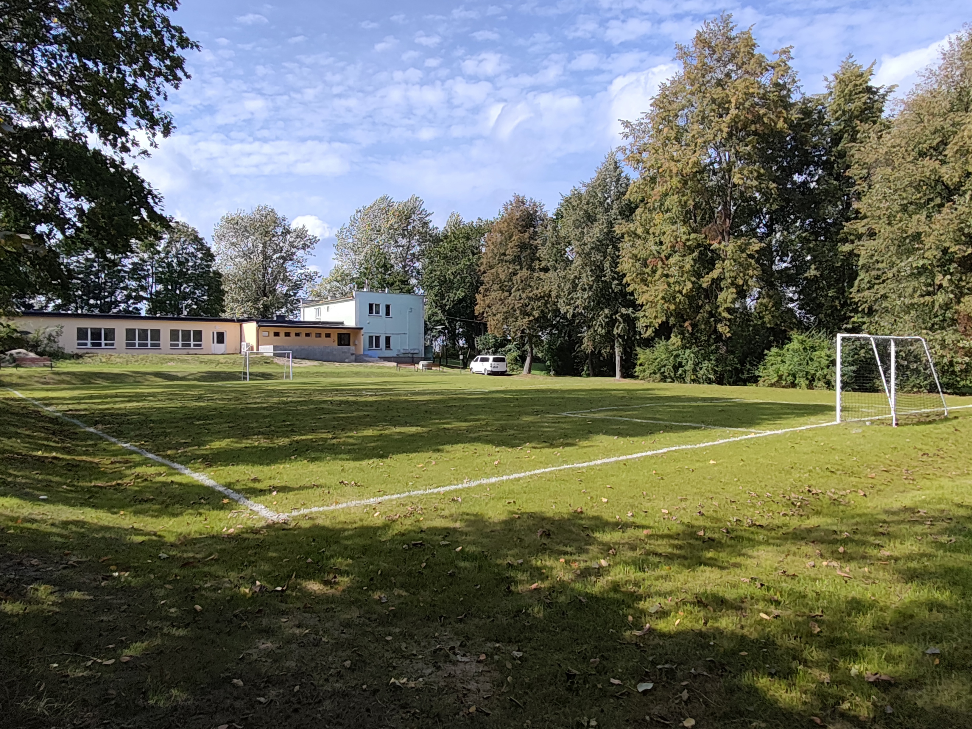 Trawiaste boisko piłkarskie z białymi liniami i dwiema bramkami. W tle znajduje się niski budynek oraz gęsto zadrzewiony teren. Na boisku nie ma zawodników. Gleba i trawa są oświetlone słońcem, a nad boiskiem widać częściowo zachmurzone niebo.