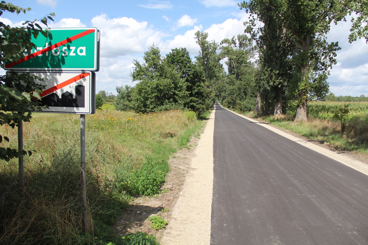 Nowy asfalt na drodze ciągnie się kilometrami. Po obu stronach białe pobocza, po lewej tablica informacyjna, w tle drzewa, łąki, lasy