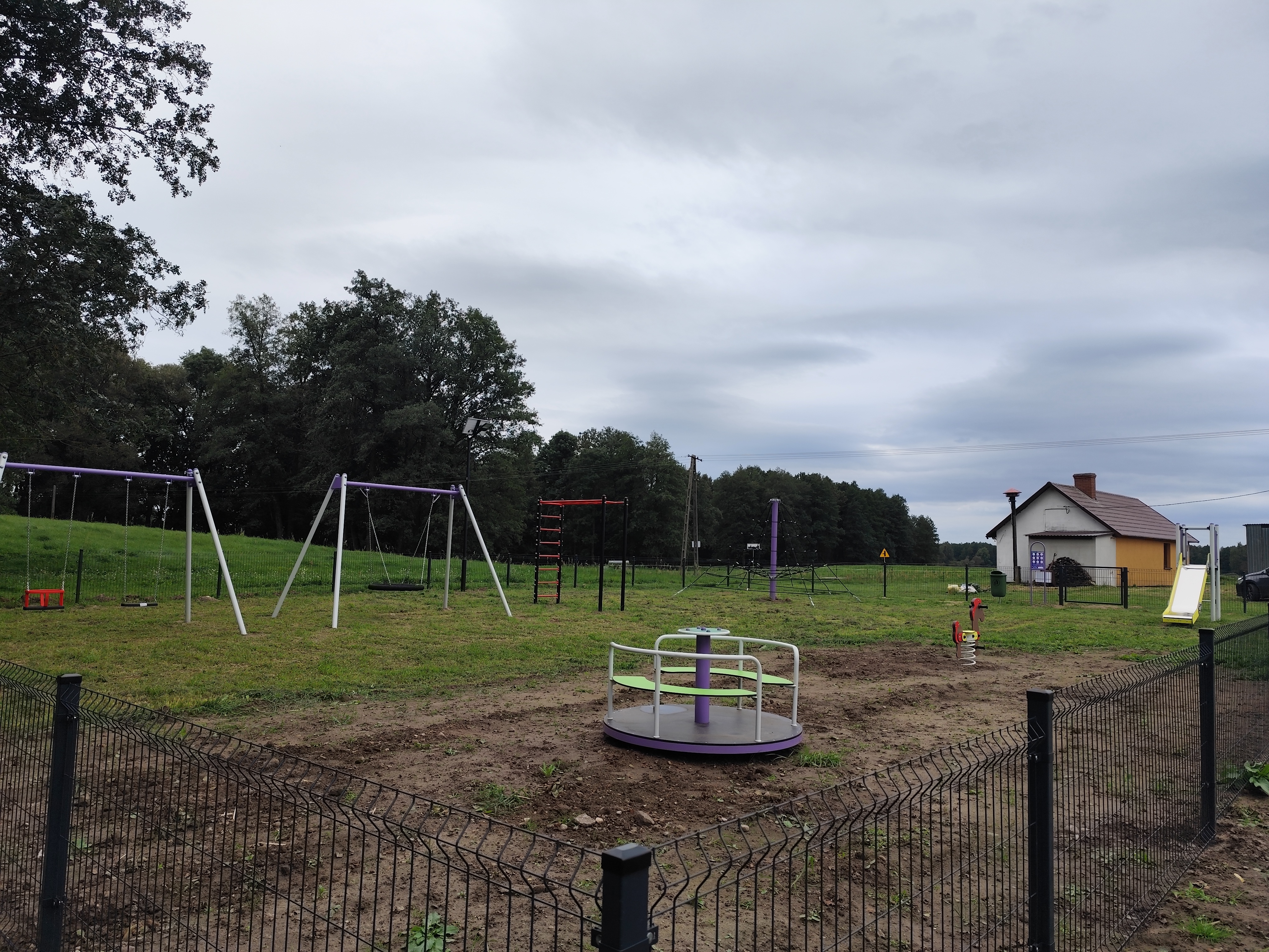Zdjęcie przedstawia wiejski plac zabaw ogrodzony metalowym płotem. Widać na nim huśtawki, karuzelę, zjeżdżalnię oraz drabinki do wspinaczki. W tle znajduje się mały dom z żółtą elewacją i czerwonym dachem, a za nim rozciąga się las i zielone pola pod pochmurnym niebem.