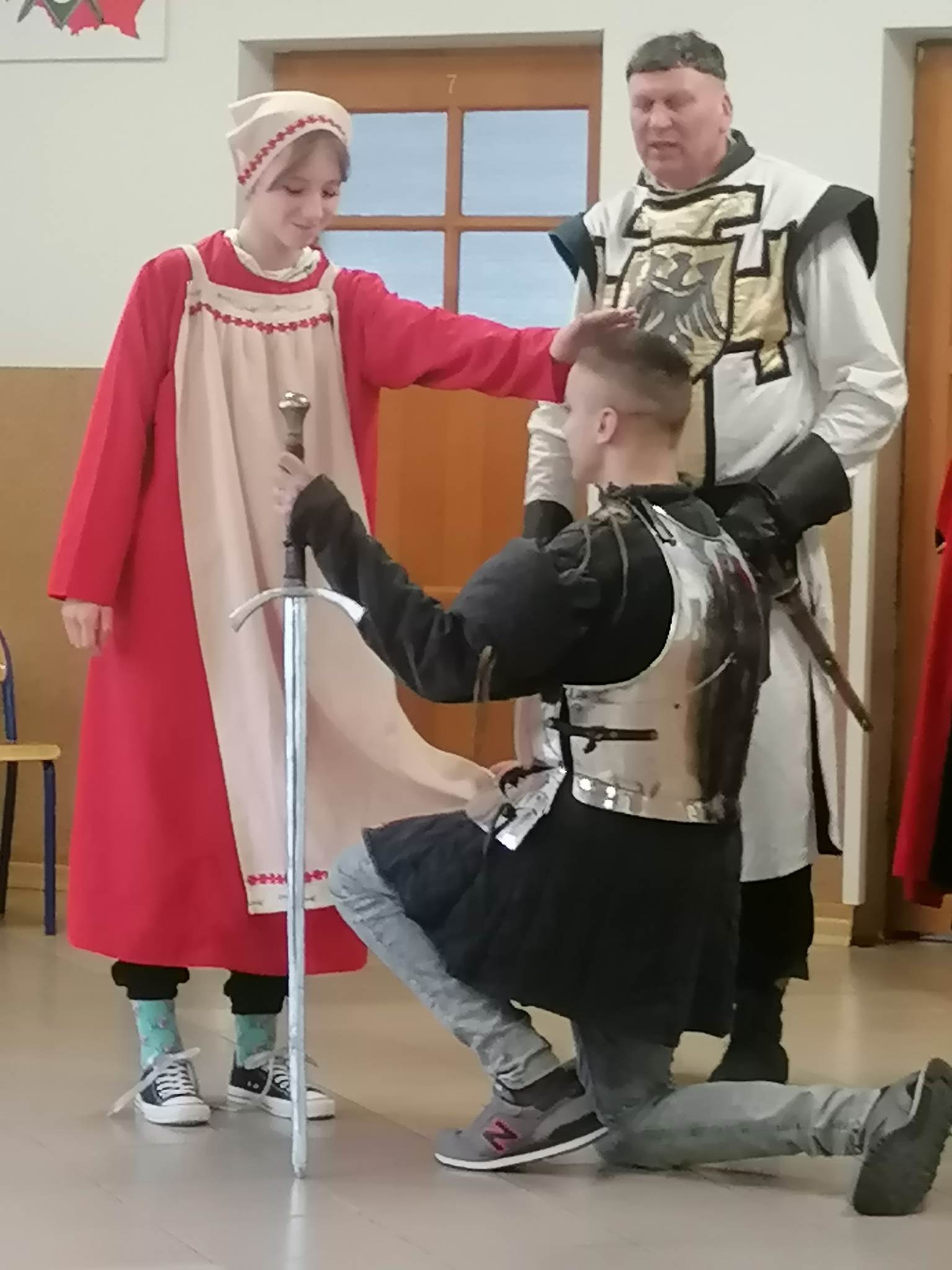 Uczennica ubrana w starodawną sukienkę z czepkiem nachyla się nad chłopakiem przebranym za rycerza.