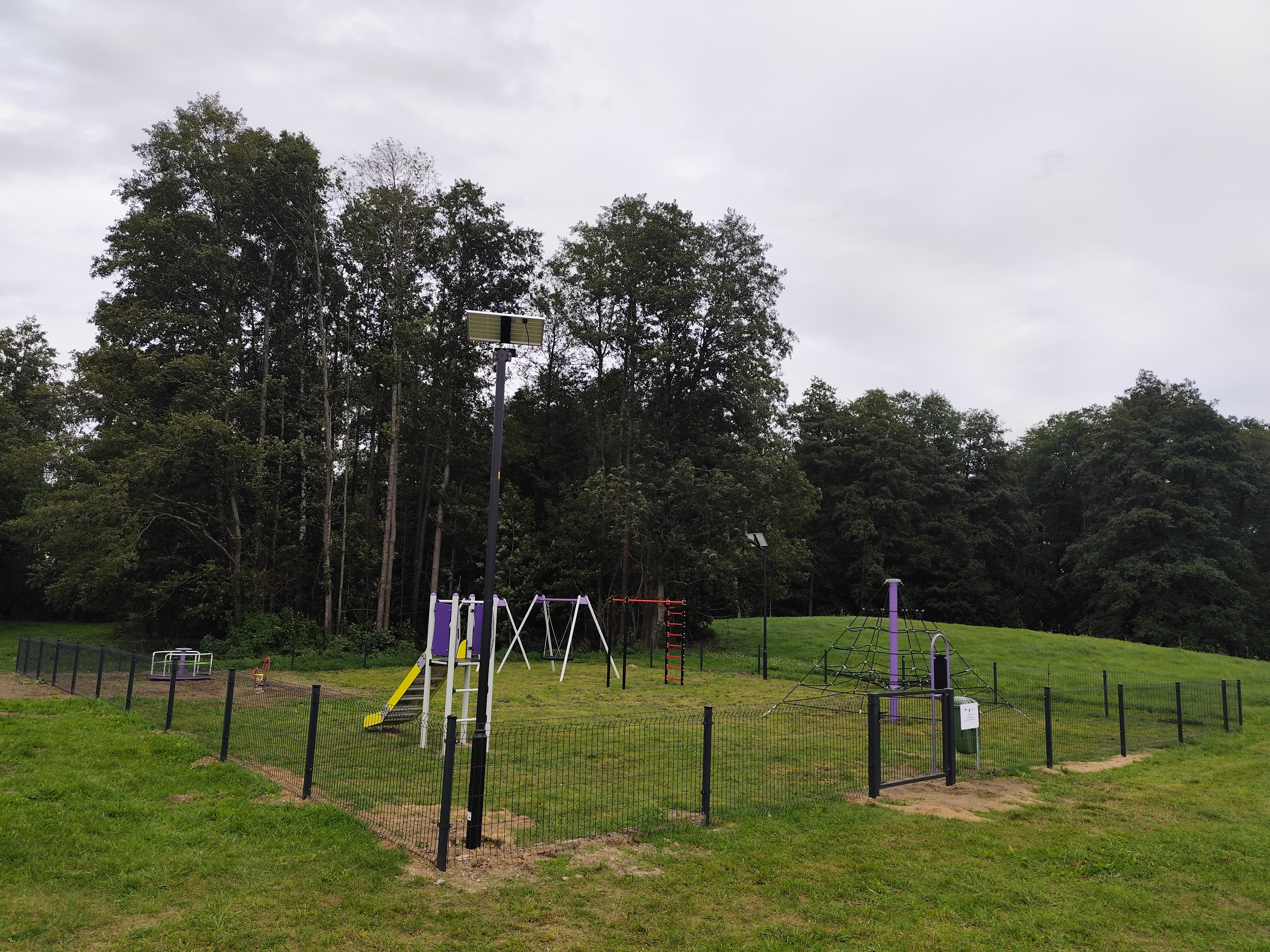 Zdjęcie przedstawia wiejski plac zabaw ogrodzony metalowym płotem. Widać na nim huśtawki, karuzelę, zjeżdżalnię oraz drabinki do wspinaczki.