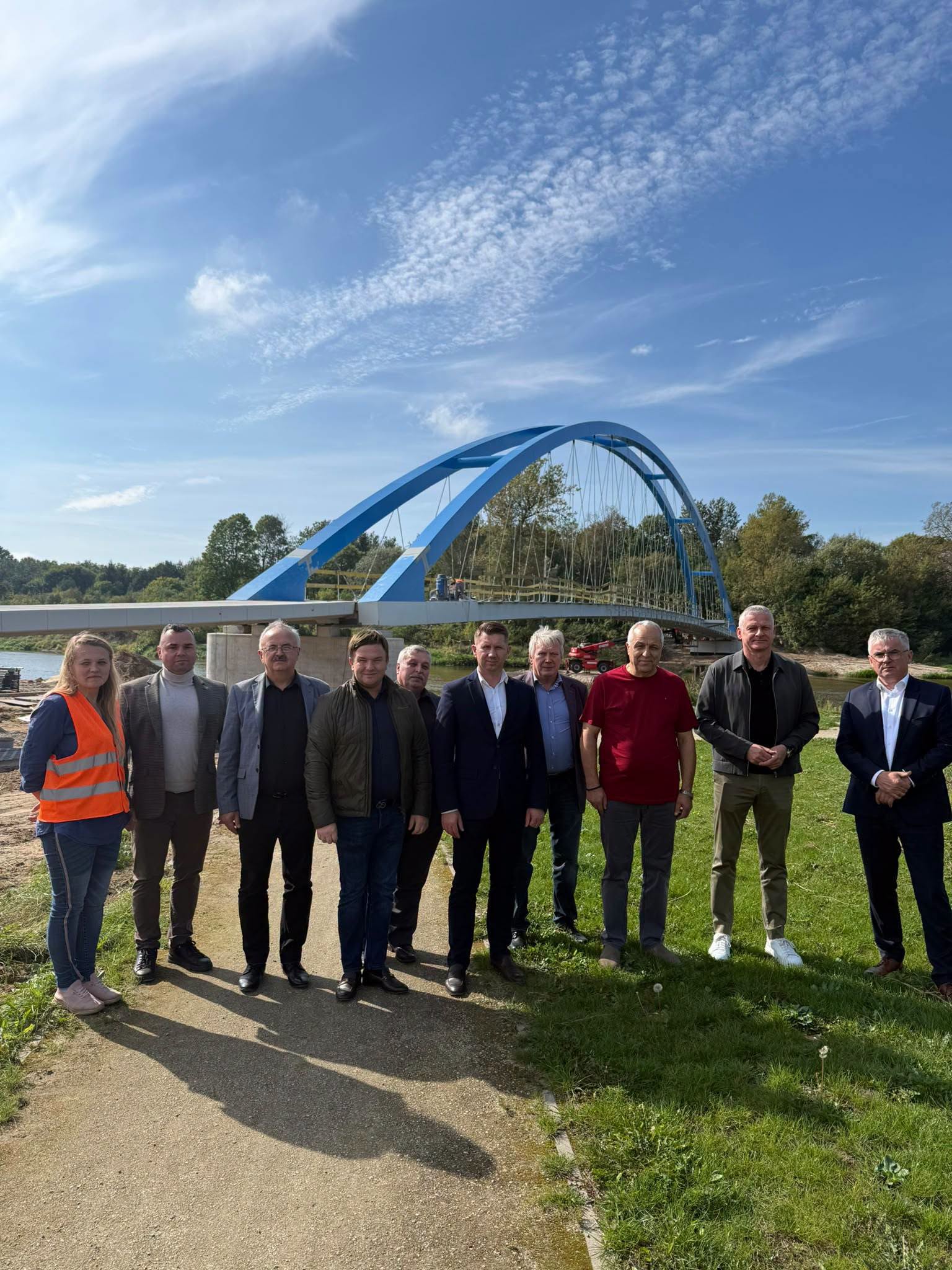 Grupa dzieisięciu osób pozuje nad rzeką, za którą znajduje się niebieski łukowy most i porośnięty drzewami krajobraz.