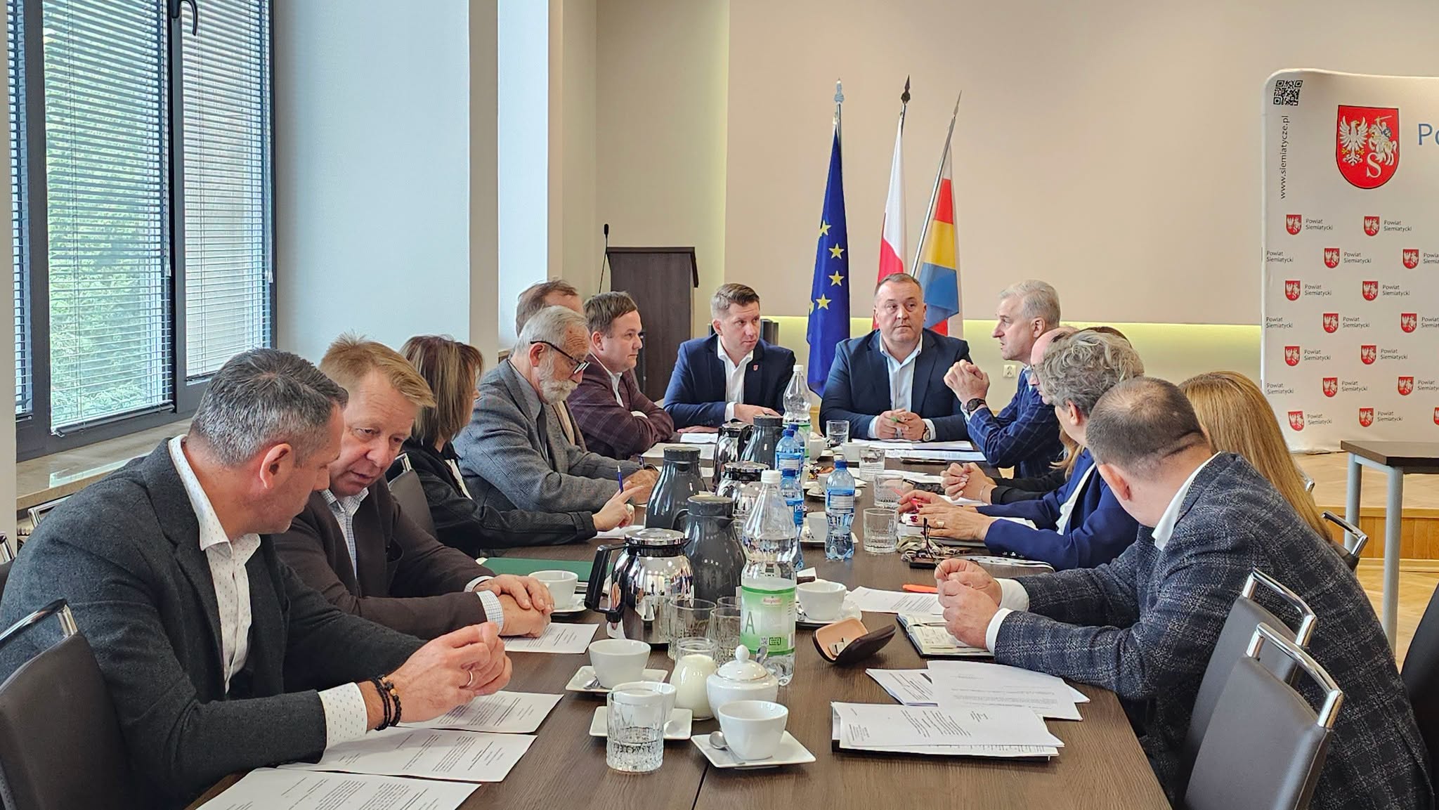 Spotkanie odbywające się przy stole konferencyjnym z udziałem różnych osób, z flagami w tle i dokumentami rozłożonymi na stole.