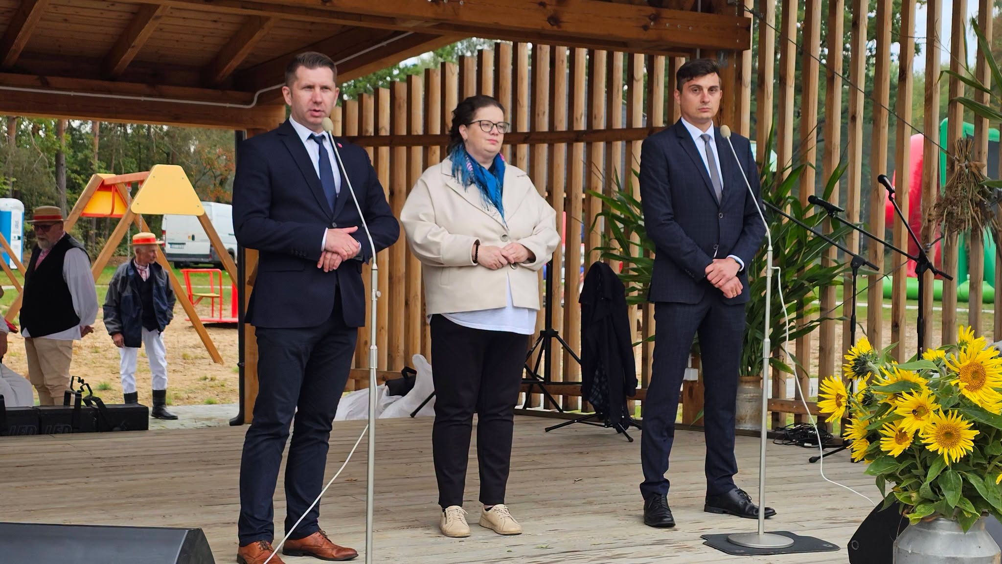 Trzy osoby stoją na scenie pod drewnianą konstrukcją: jedna kobieta w jasnym płaszczu i dwóch mężczyzn w ciemnych garniturach. Na scenie znajdują się rośliny i słoneczniki, a w tle widać kolorowe urządzenia na placu zabaw.