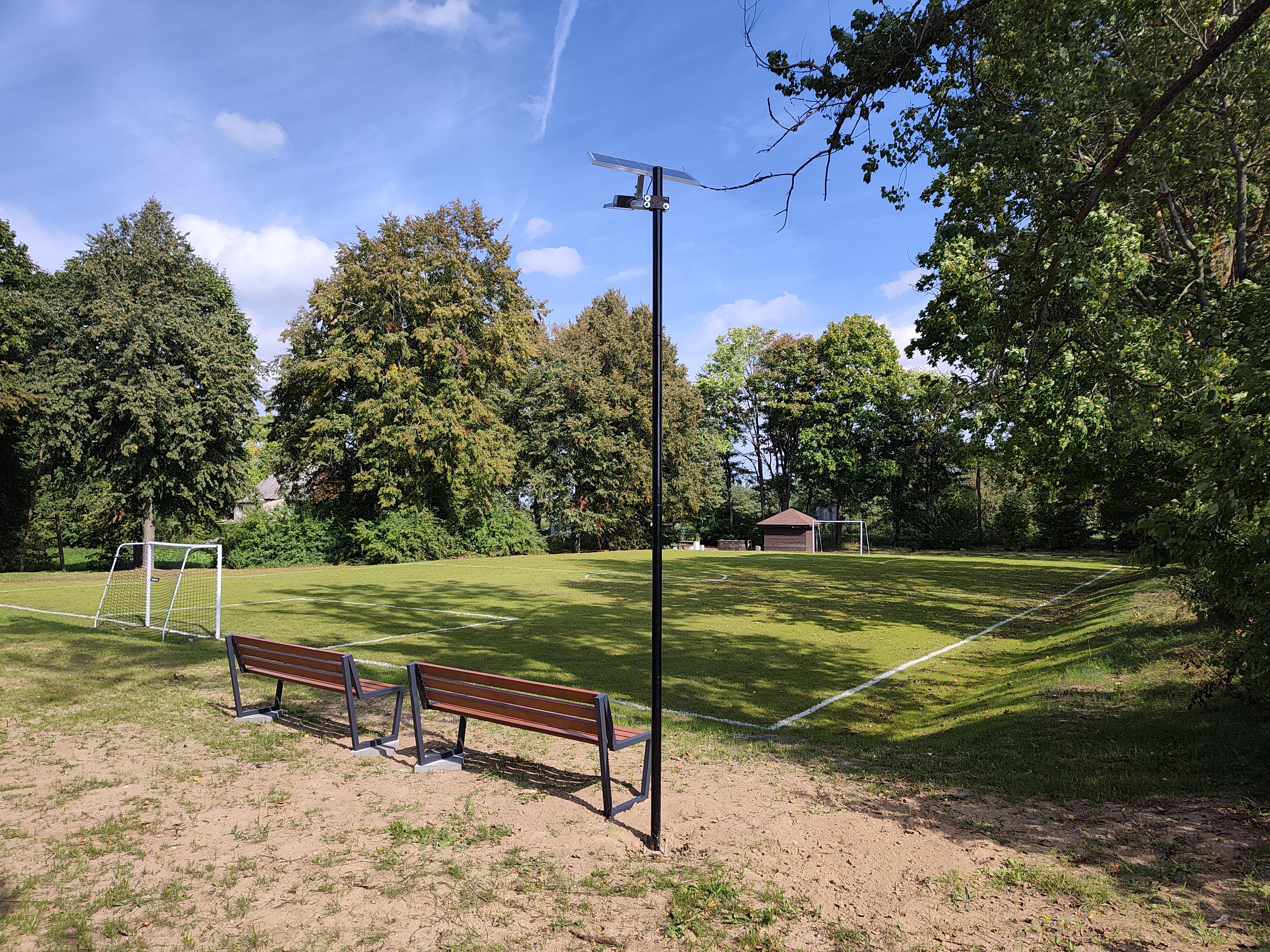 Trawiaste boisko piłkarskie z dwiema bramkami i białymi liniami. Wzdłuż boiska stoją dwie drewniane ławki, a pośrodku znajduje się wysoka lampa solarna. W tle rosną drzewa i krzewy. Nad boiskiem widać błękitne niebo z chmurami.