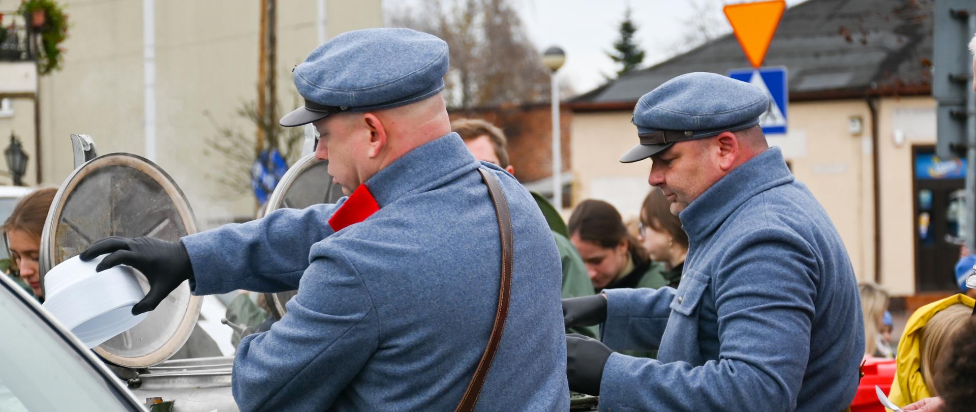 Na zdjęciu widzimy trzech kucharzy w strojach wojskowych, którzy przygotowują i wydają grochówkę z kuchni polowej. Mężczyźni są zaangażowani w pracę, a ich stroje wojskowe dodają powagi i profesjonalizmu. Widać, że są dobrze zorganizowani i skoncentrowani na swoim zadaniu. 