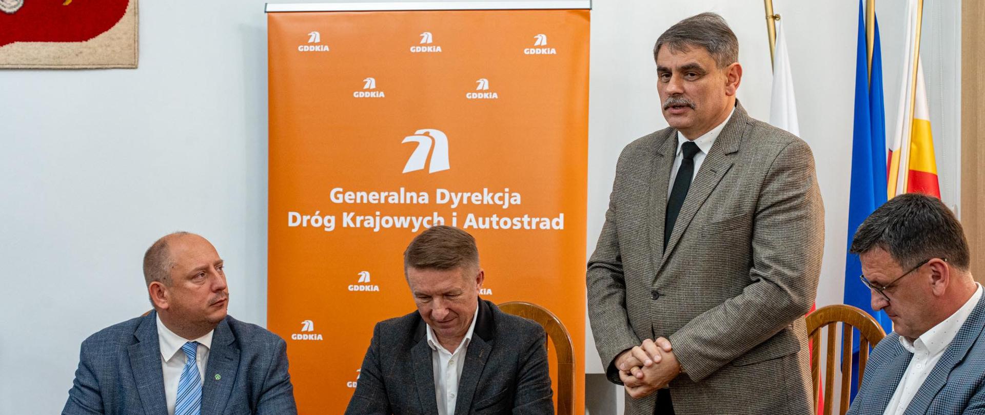 Na zdjęciu czterech mężczyzn siedzi i stoi przy stole podczas oficjalnego spotkania. Po prawej stronie stoi Burmistrz Kalwarii Zebrzydowskiej Tadeusz Stela, przemawiając w kierunku pozostałych uczestników. Za nim widoczne są flagi – polska, unijna oraz województwa małopolskiego. W tle stoi pomarańczowy roll-up z logo i nazwą „Generalna Dyrekcja Dróg Krajowych i Autostrad”. Po lewej stronie kadru znajduje się fragment godła Polski. Mężczyźni mają na sobie garnitury i wyglądają na zaangażowanych w oficjalne podpisanie dokumentów lub rozmowę służbową.