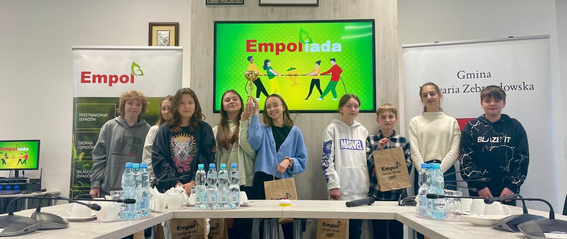 W jasnej, oświetlonej sali stoją za stolikami uśmiechnięci młodzi ludzie, trzymając papierowe torby z nagrodami i puchar; na stołach woda w małych butelkach i mikrofony, w tle rollupy organizatorów, na ścianie obrazy, godło i krzyż