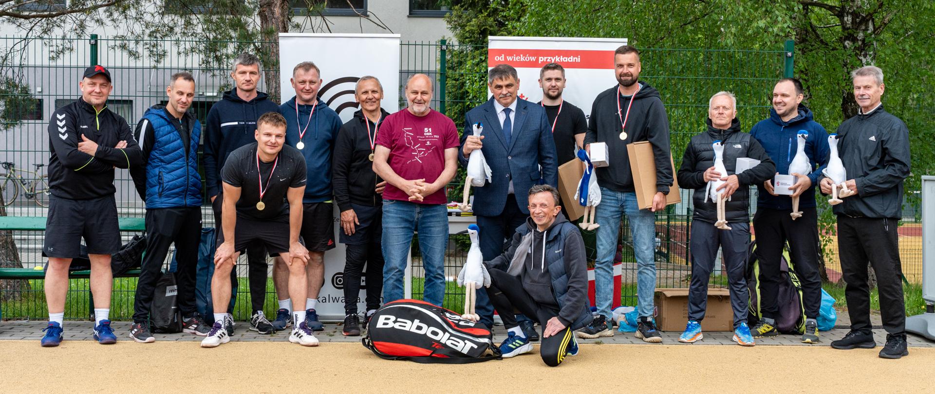Grupowe zdjęcie uczestników II Turnieju Deblowego Tenisa Ziemnego o Puchar Doliny Karpia wykonane na korcie tenisowym w Kalwarii Zebrzydowskiej. Na fotografii widoczni są zawodnicy, organizatorzy oraz przedstawiciele samorządu stojący i klęczący na tle ogrodzenia oraz zieleni. Część uczestników ma na szyjach medale, a kilku trzyma puchary oraz nagrody. Na pierwszym planie znajduje się torba tenisowa marki Babolat. Zdjęcie wykonano po zakończeniu turnieju podczas wspólnej pamiątkowej fotografii.
