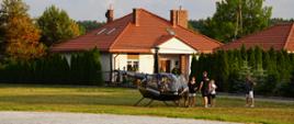 Na zdjęciu widać czarny helikopter na tle domu jednorodzinnego, który wylądował na trawniku. Z helikoptera wysiada grupa ludzi. 
