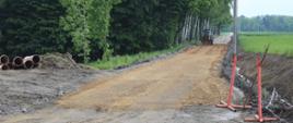 Droga Wudyka pozbawiona asfaltowej nawierzchni. Po lewej las i rury instalacyjne, po prawej łąka. W oddali koparka na drodze.