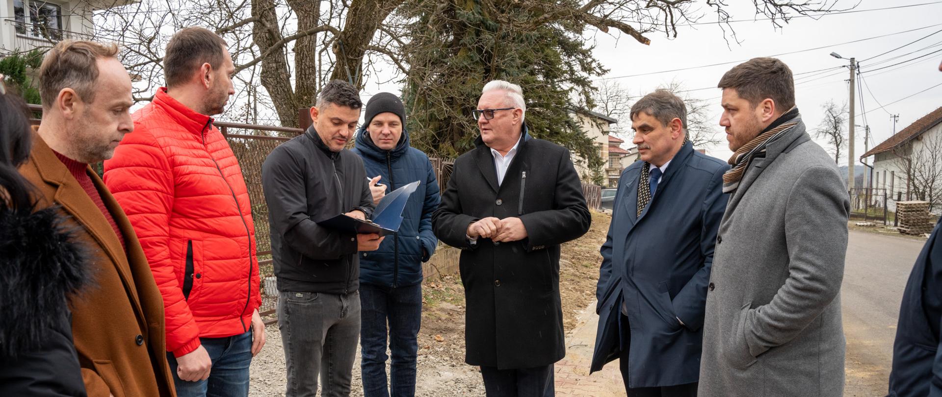 Grupa przedstawicieli samorządu i pracowników technicznych stoi przy nowo układanym chodniku wzdłuż drogi lokalnej. Jeden z mężczyzn trzyma dokumentację i omawia szczegóły inwestycji, pozostali słuchają i obserwują zakres prac. Spotkanie odbywa się na zewnątrz, w terenie zabudowy jednorodzinnej, w okresie zimowym.