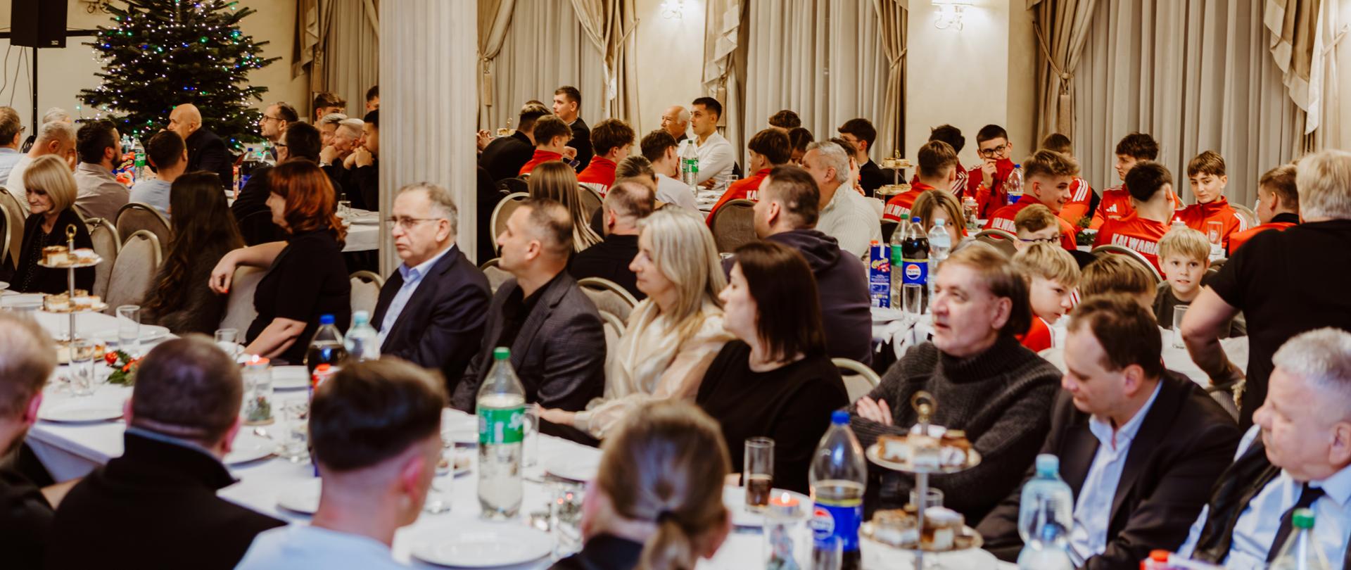 Uczestnicy spotkania wigilijnego siedzą przy długich stołach w eleganckiej, jasnej sali z kolumnami i żyrandolami. W tle widoczna jest udekorowana choinka bożonarodzeniowa. Przy stołach zasiadają mieszkańcy oraz młodzi sportowcy w czerwonych strojach klubowych, rozmawiający i wspólnie spędzający czas przy świątecznym poczęstunku.